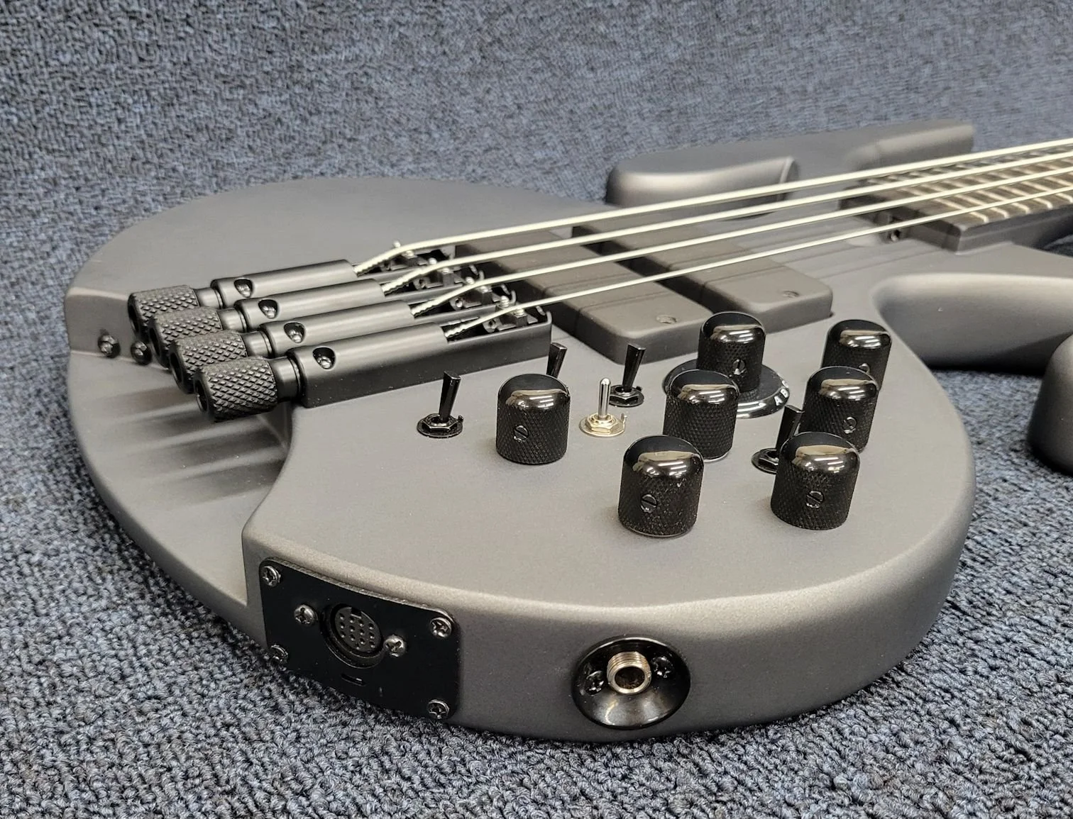 Close-up of a sleek, modern electric bass guitar with a grey body, black control knobs, bridge, and input jack, resting on a textured grey fabric surface.