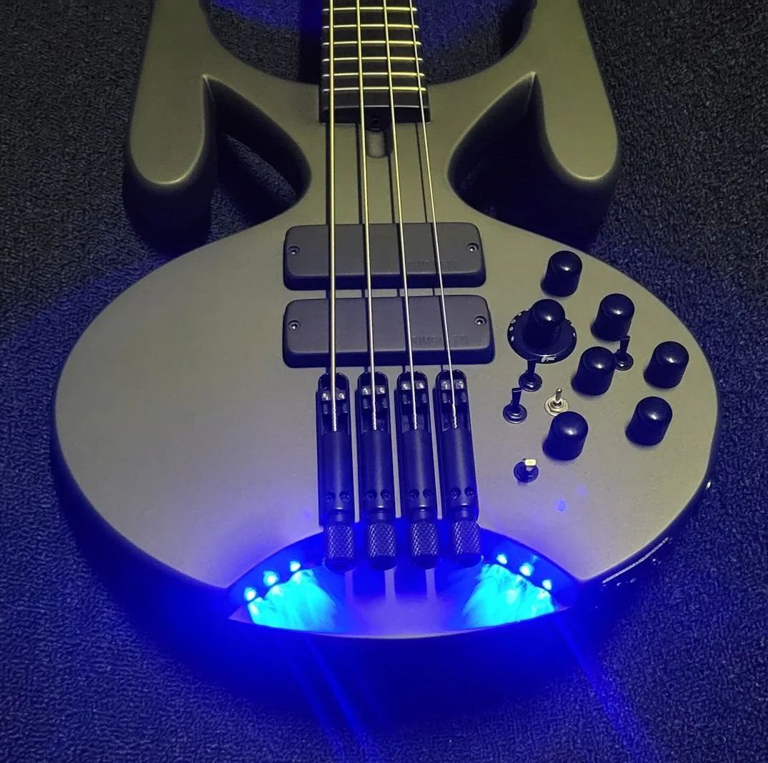 Black electric bass guitar with four strings, control knobs, and blue LED lights at the bottom, resting on a textured black surface.