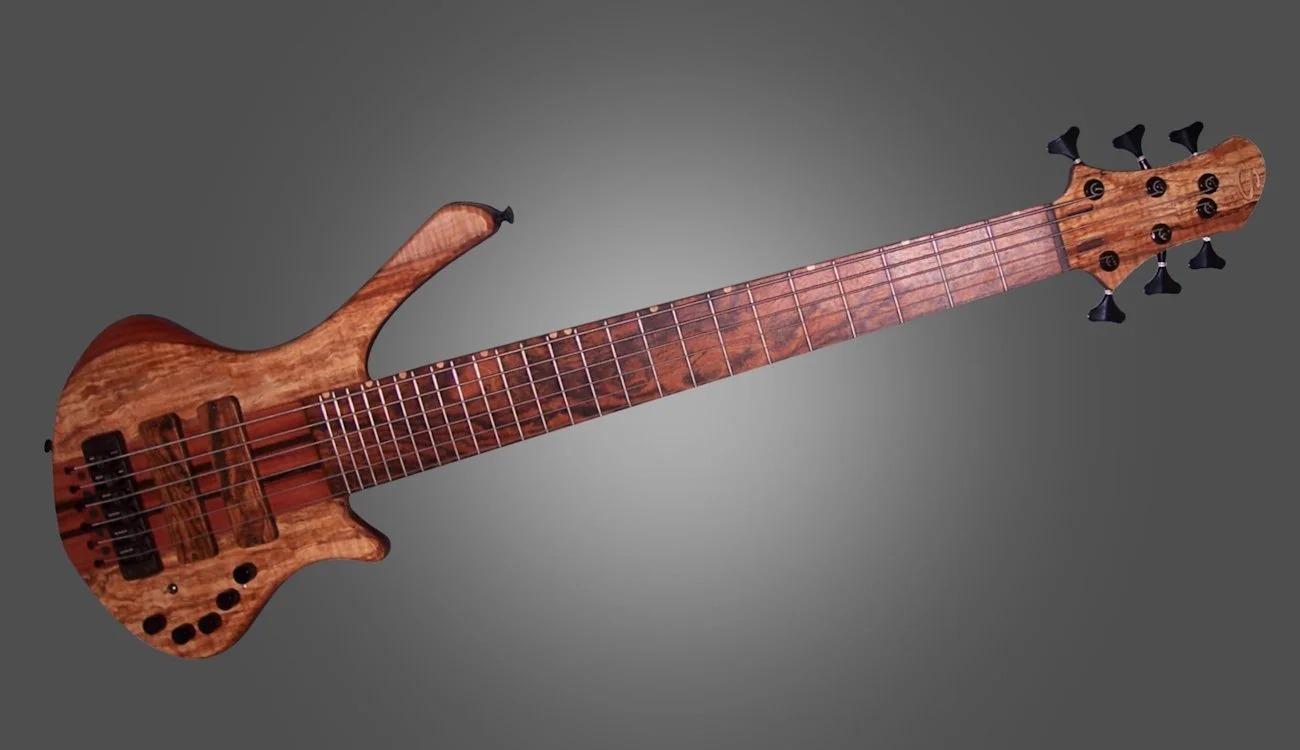 A wooden electric bass guitar with six strings, multiple knobs, and tuning pegs, set against a gray background.