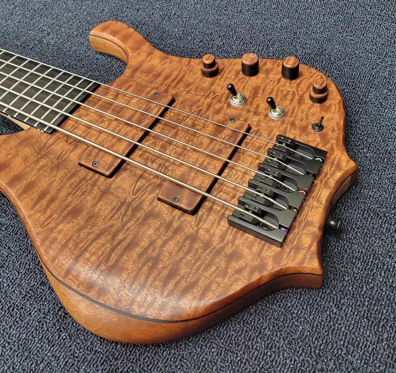 A wooden electric bass guitar with black hardware and a quilted wood grain finish, resting on a black textured surface.