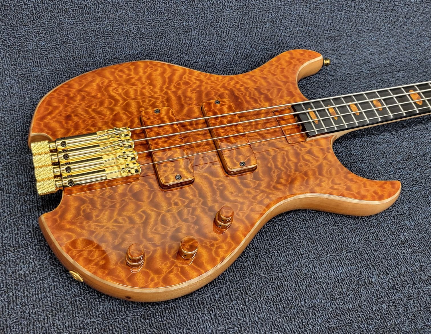 A close-up of a handmade electric bass guitar with a quilted maple wood finish and gold hardware, resting on a dark textured surface.