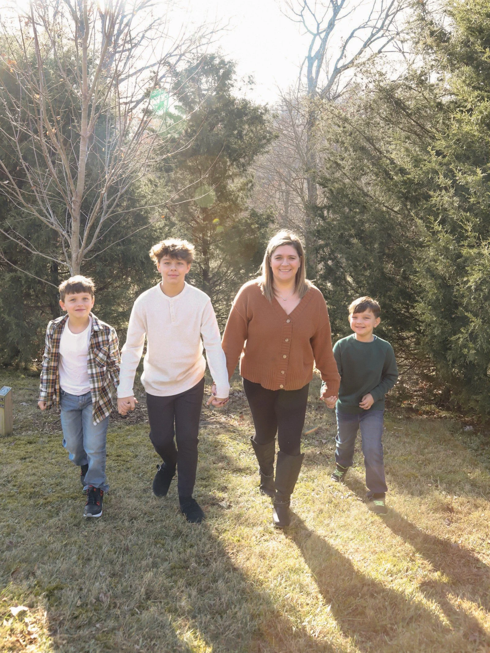 A group of five people, a woman and four boys, walking together outdoors on a sunny day through a grassy area with trees in the background.
