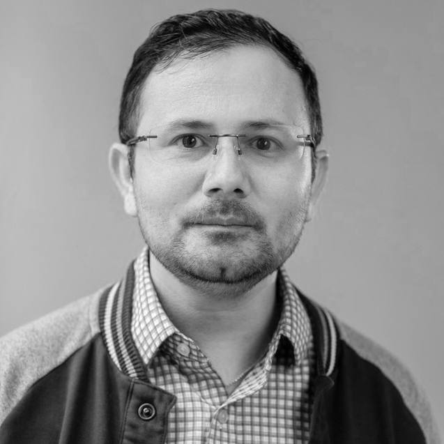 Black and white portrait of a man wearing glasses, a checkered shirt, and a varsity jacket, looking directly at the camera with a neutral expression.
