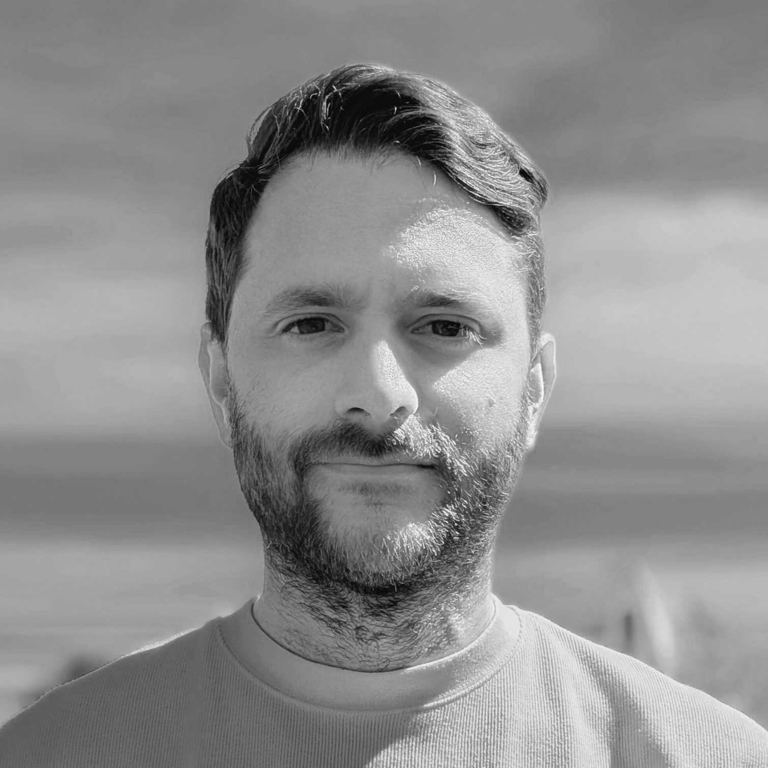 Black and white photo of a young man with short hair and a beard, standing outdoors with a cloudy sky in the background.