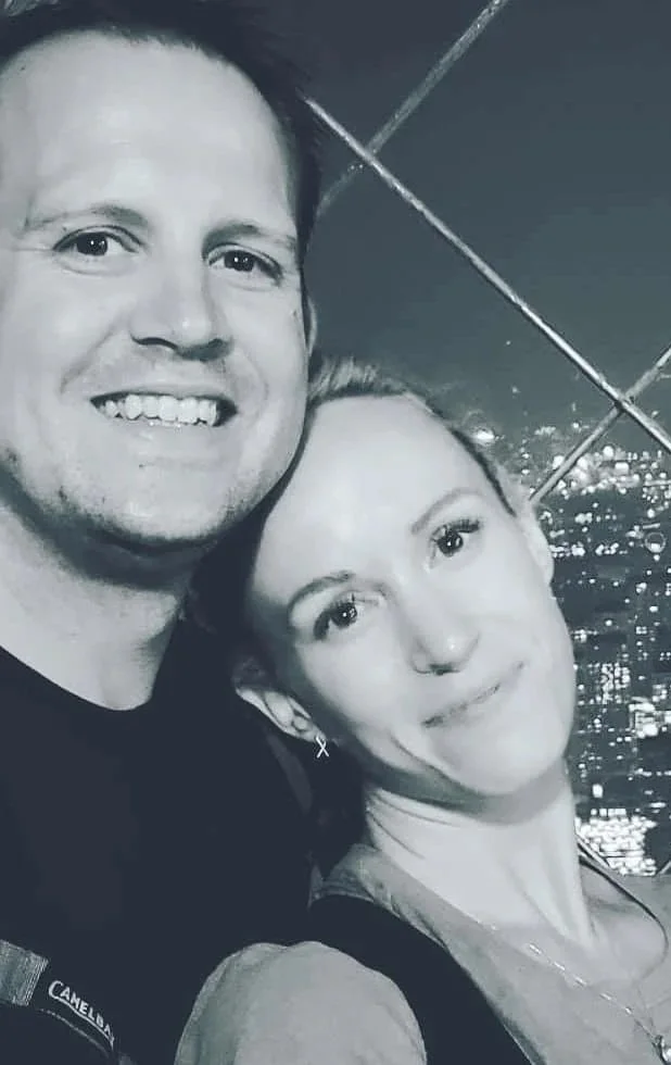 A black-and-white photo of a smiling man and woman close together, with a cityscape and lights in the background, taken at night.