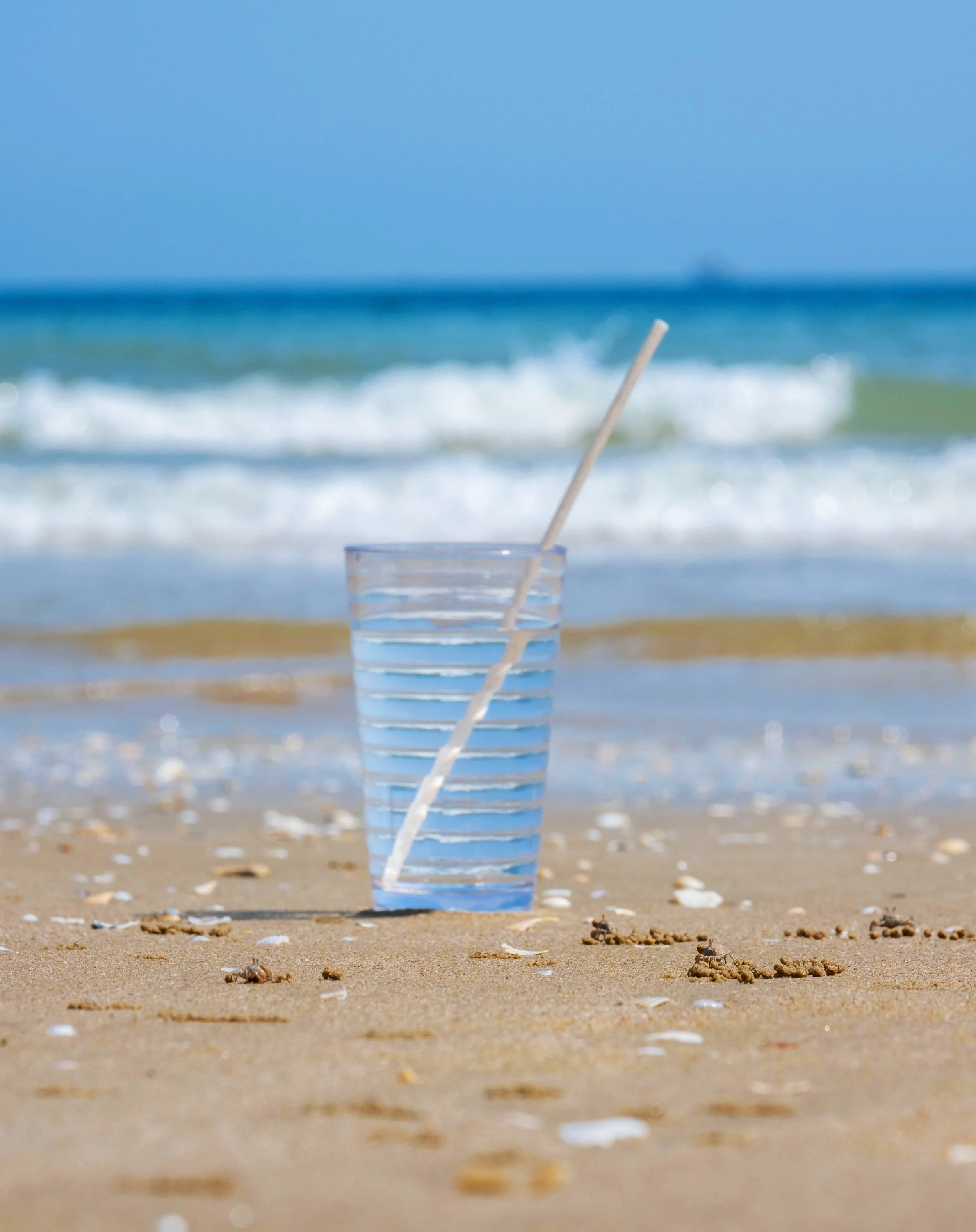 Solinatra Straw in a glass filled with water on the sandy beach near the ocean shoreline.