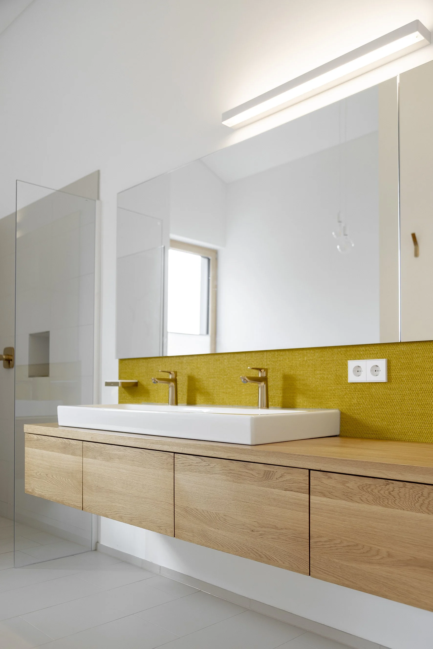 Moderner Waschraum mit Doppelwaschbecken, Holzschrank, großen Spiegeln und weißen Wänden. Es gibt eine helle, natürliche Beleuchtung durch ein Fenster.