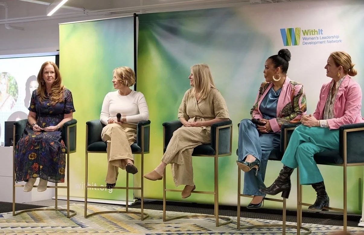 Panel of five women seated on stage during a professional leadership event with a green and blue branded backdrop.