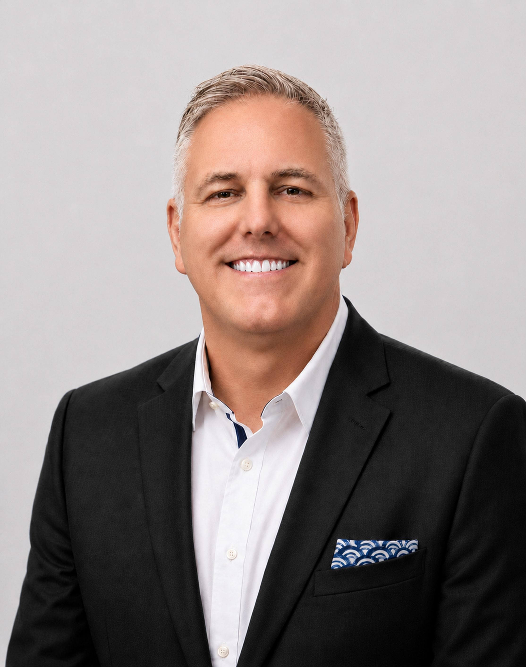 Professional headshot of a smiling man with short gray hair, wearing a black suit jacket and white dress shirt against a light gray background.