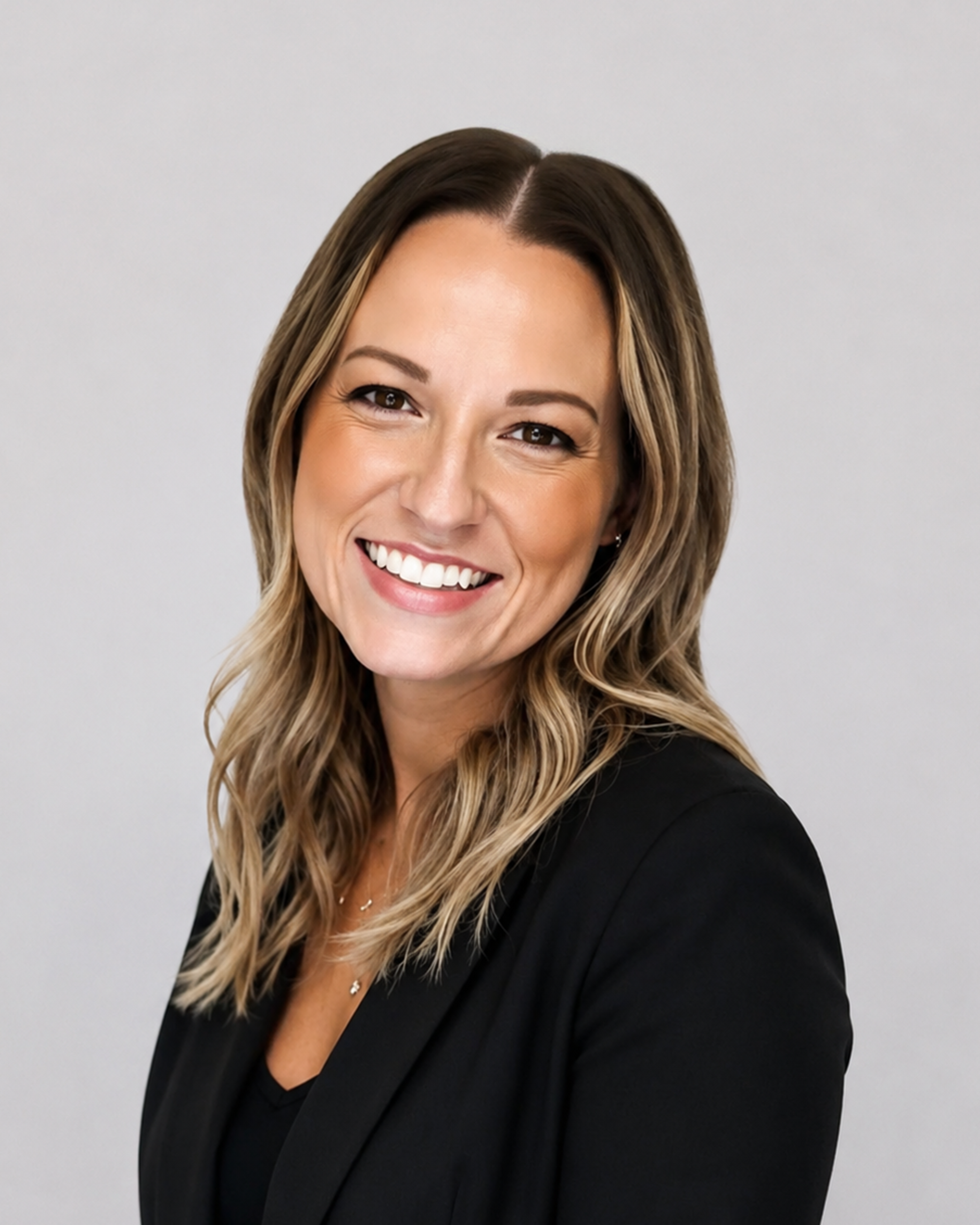 Professional headshot of a smiling woman with long highlighted brown hair, wearing a black blazer against a light gray background.