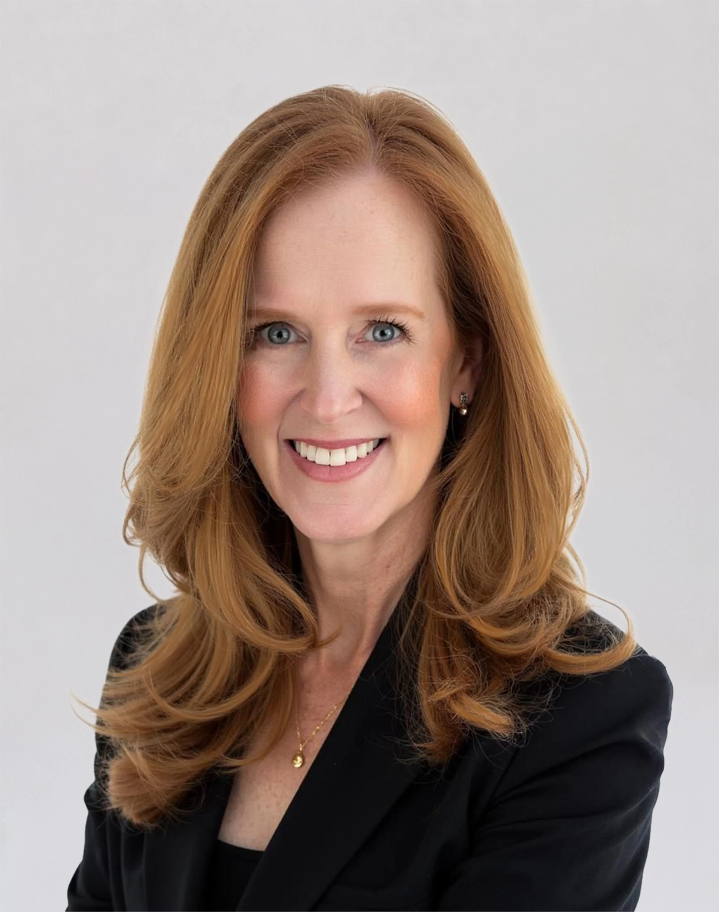 Professional headshot of a smiling woman with long auburn hair, wearing a black blazer against a light gray background.