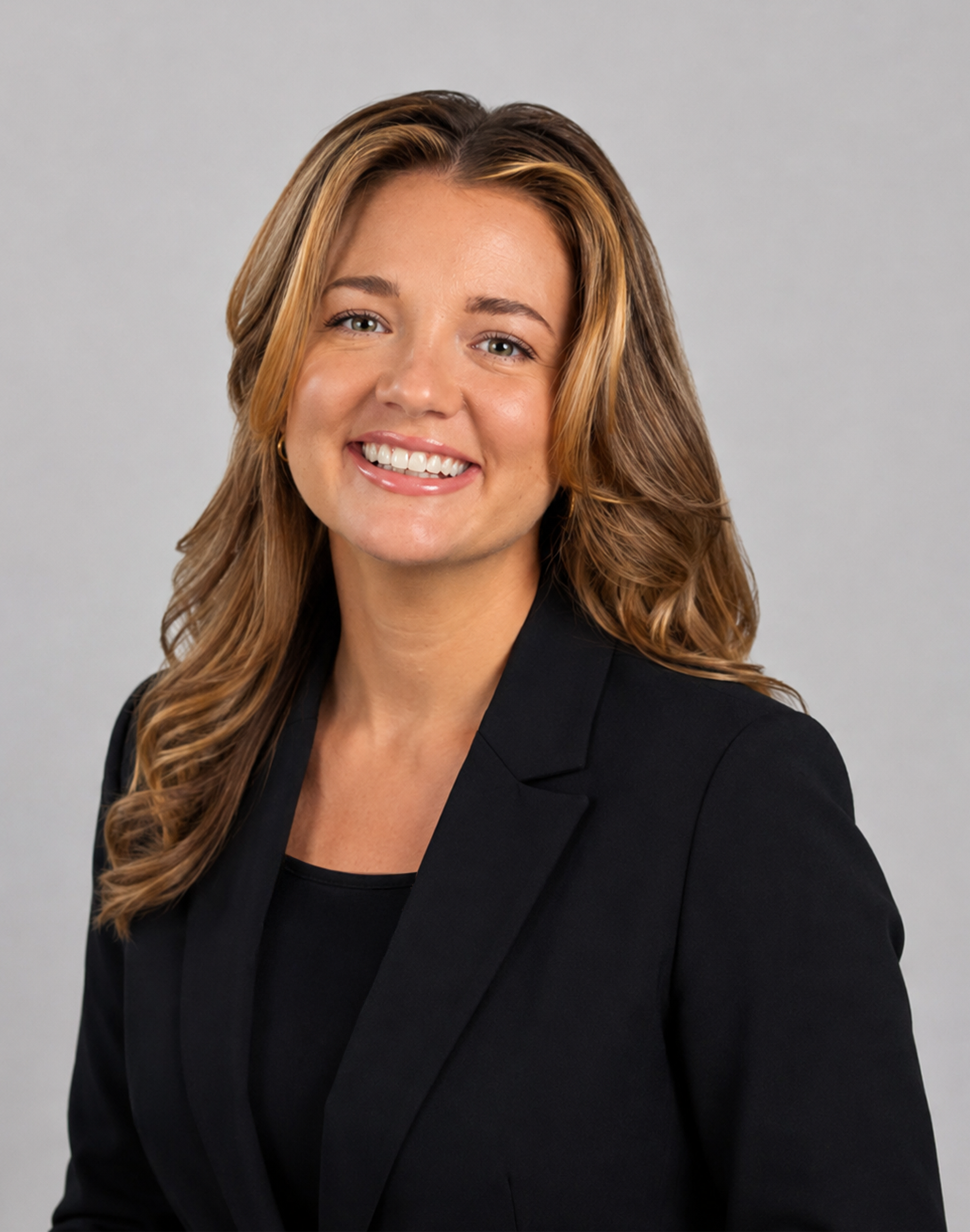 Professional headshot of a smiling woman with long light brown hair, wearing a black blazer and black top against a light gray background.