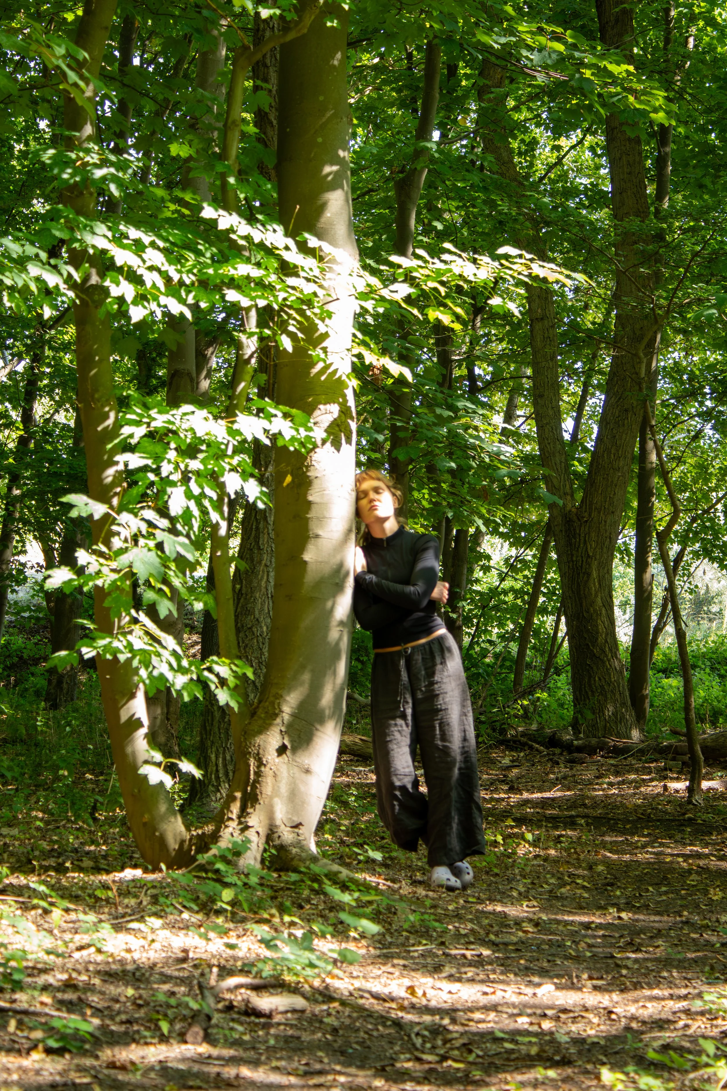 A woman in black clothing and large gray Crocs shoes standing in a forest among tall trees with green leaves, partially shaded by sunlight.