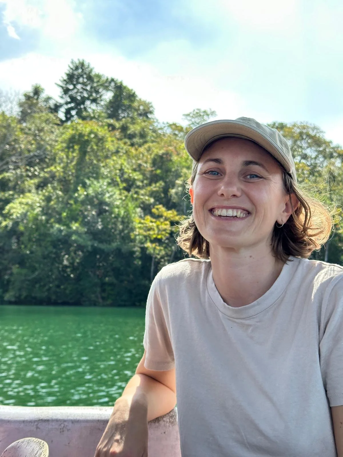 A smiling woman wearing a beige baseball cap and a light-colored t-shirt, sitting outdoors near a body of water with green trees and a partly cloudy blue sky in the background.