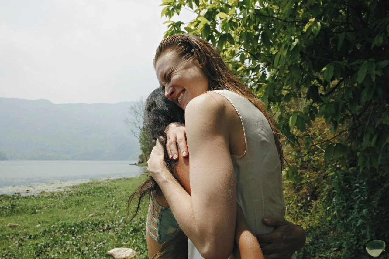 Two women hugging outdoors near a lake with green trees and mountains in the background. One woman has long brown hair and is smiling with her eyes closed, embracing the other woman.