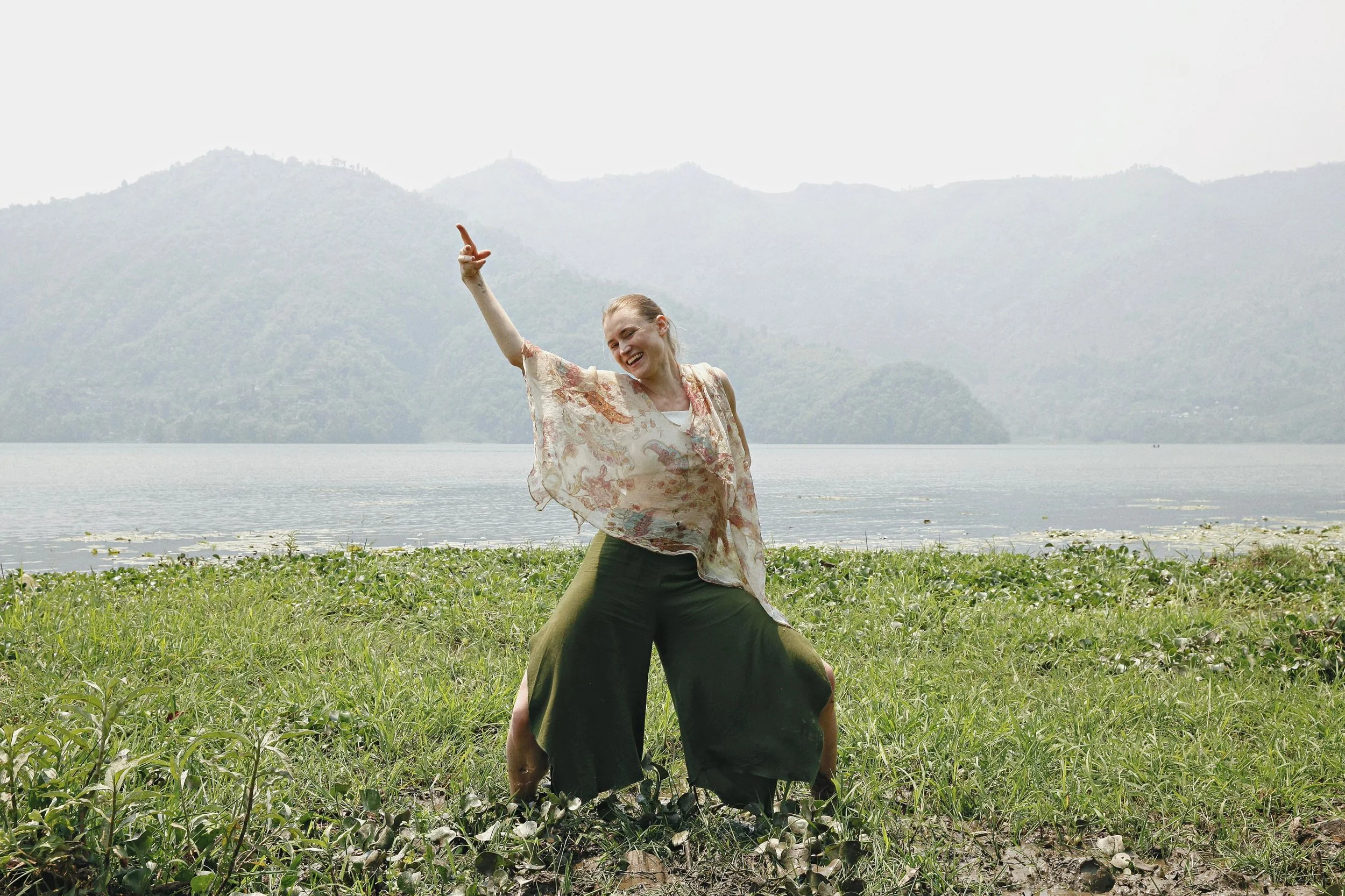 A woman standing outdoors by a lake and mountains, smiling, with the lower part of her body distorted due to the camera angle.