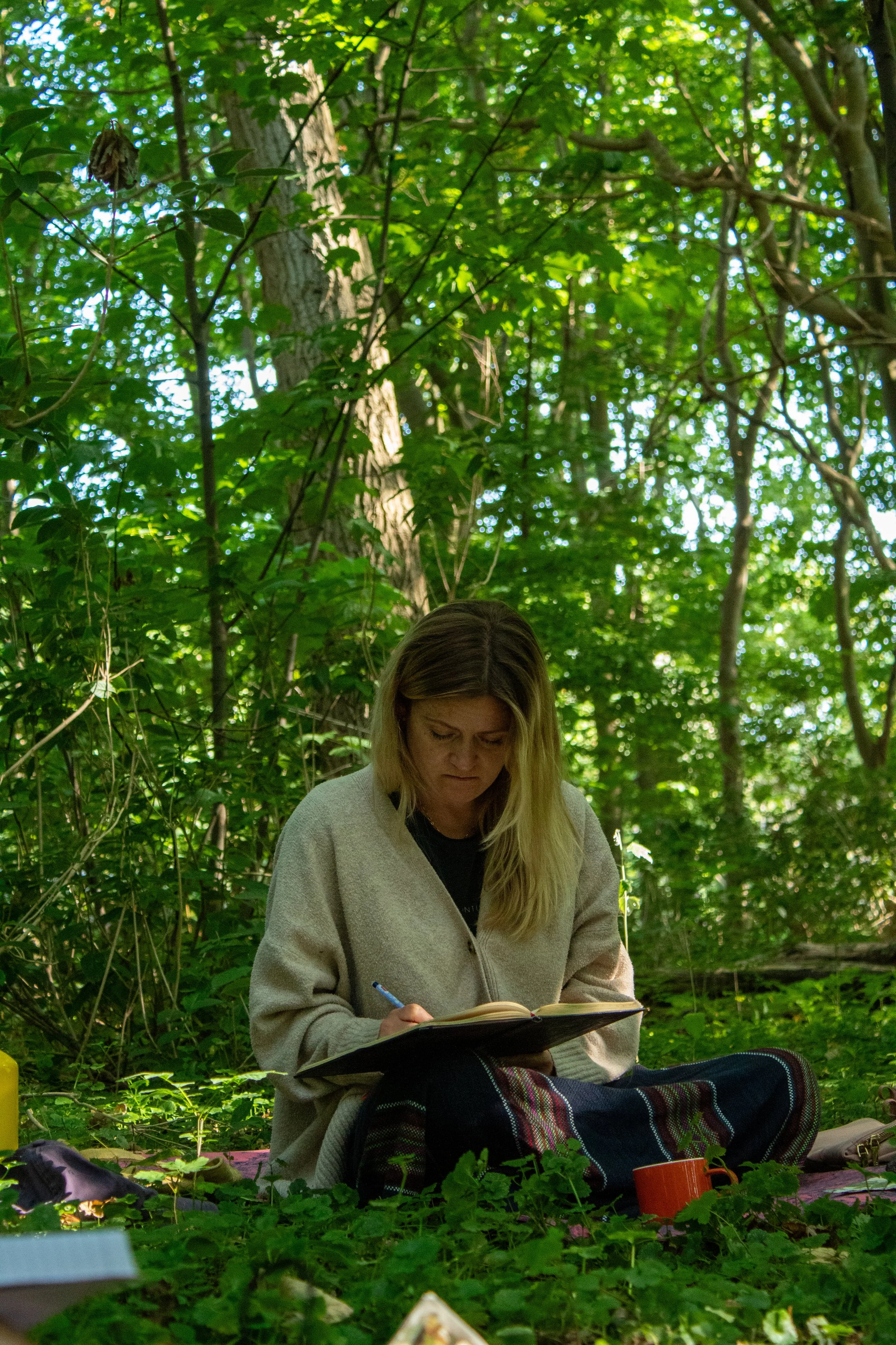 A woman with blonde hair wearing a beige jacket sitting outdoors in a wooded area, reading and taking notes from a notebook.