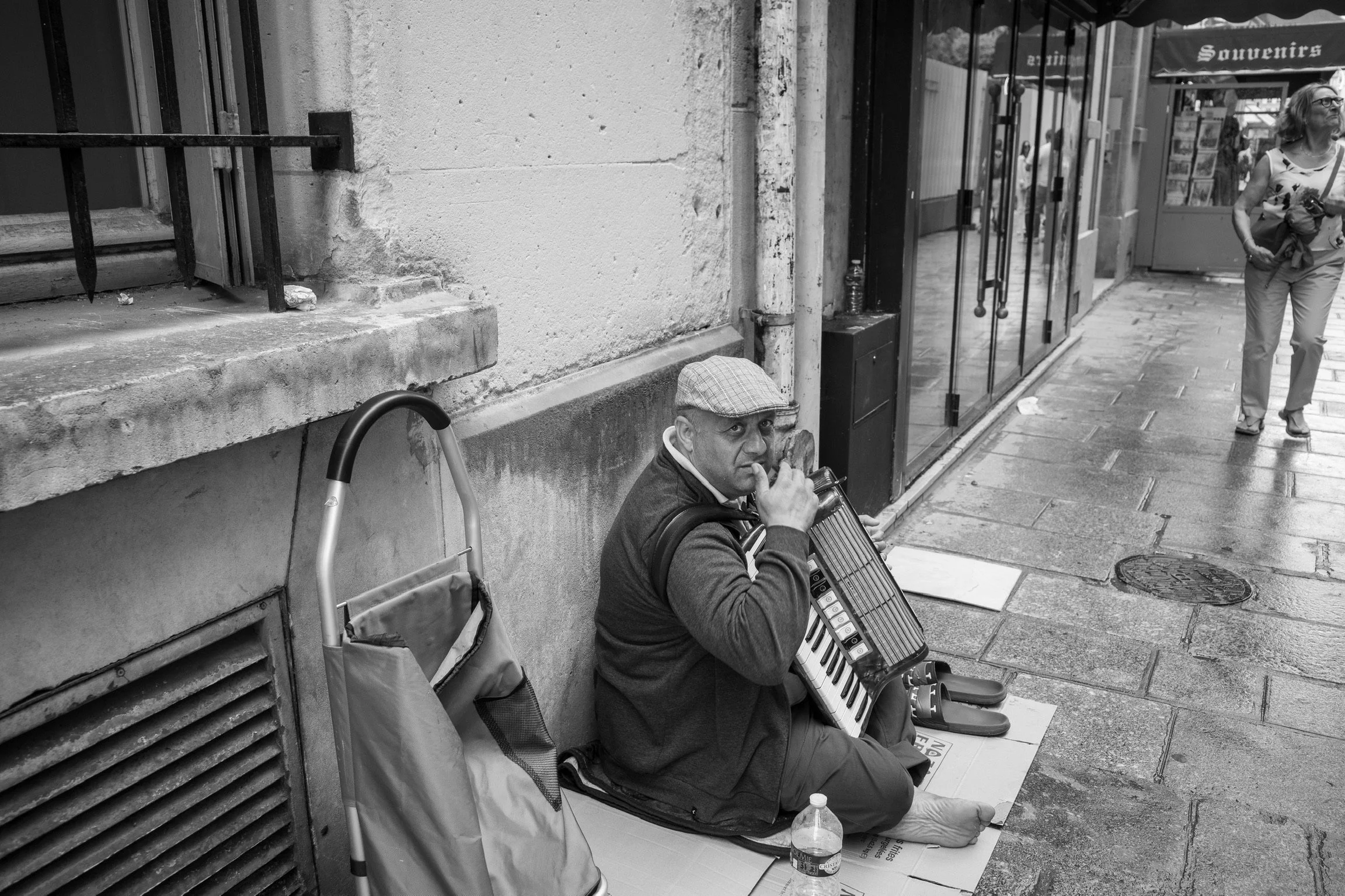 Straßenmusiker spielt auf einem Melodica an einer Hauswand sitzend. Eine Frau passiert ihn auf dem Bürgersteig. Schwarz-weiß Aufnahme.