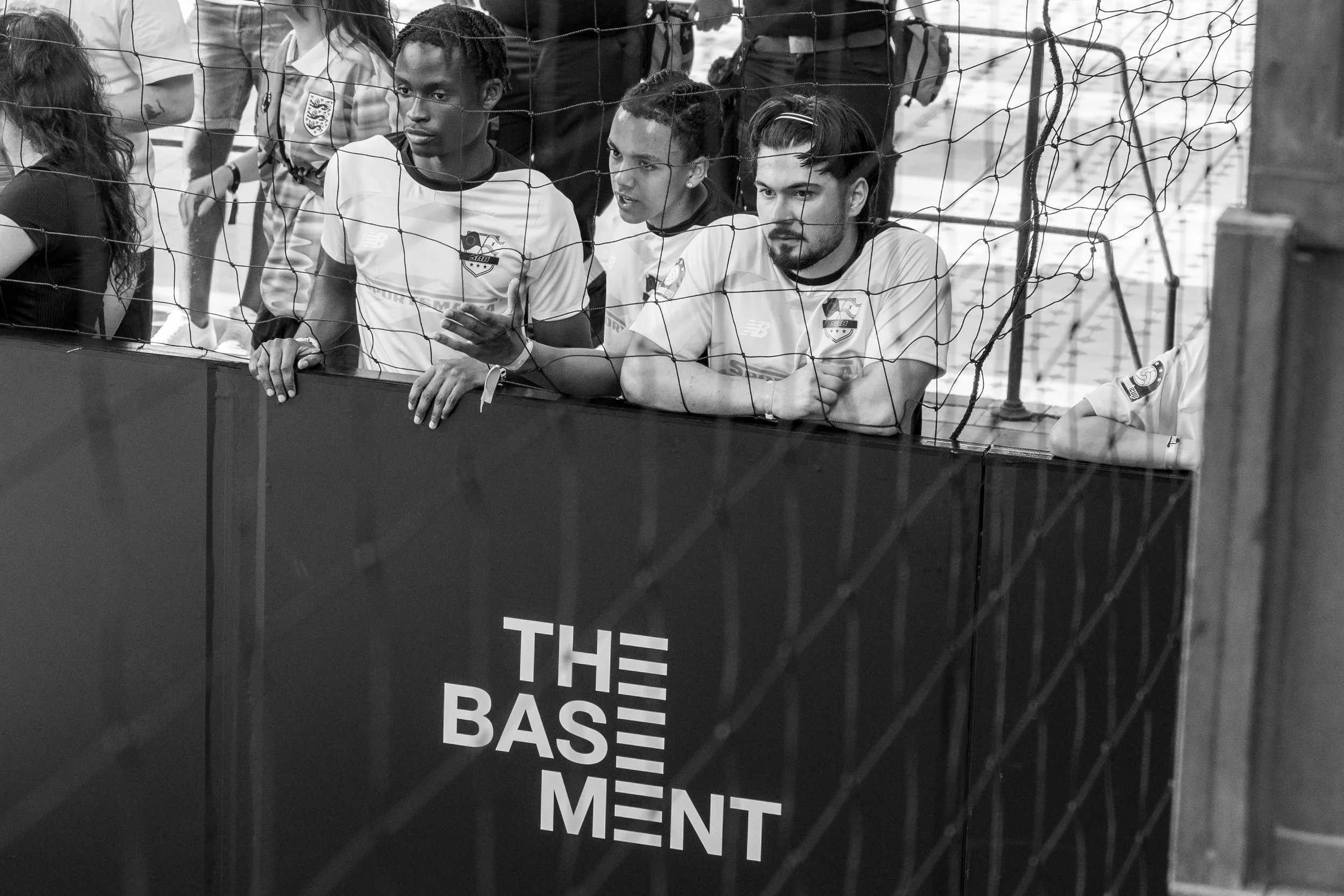 Drei junge Männer in weißen Sporttrikots stehen hinter einem Zaun und lehnen sich darauf. Auf dem Zaun ist der Schriftzug "THE BASEMENT" zu sehen. Im Hintergrund sind weitere Menschen und ein Spielfeld erkennbar. Schwarz-weiß Aufnahme.