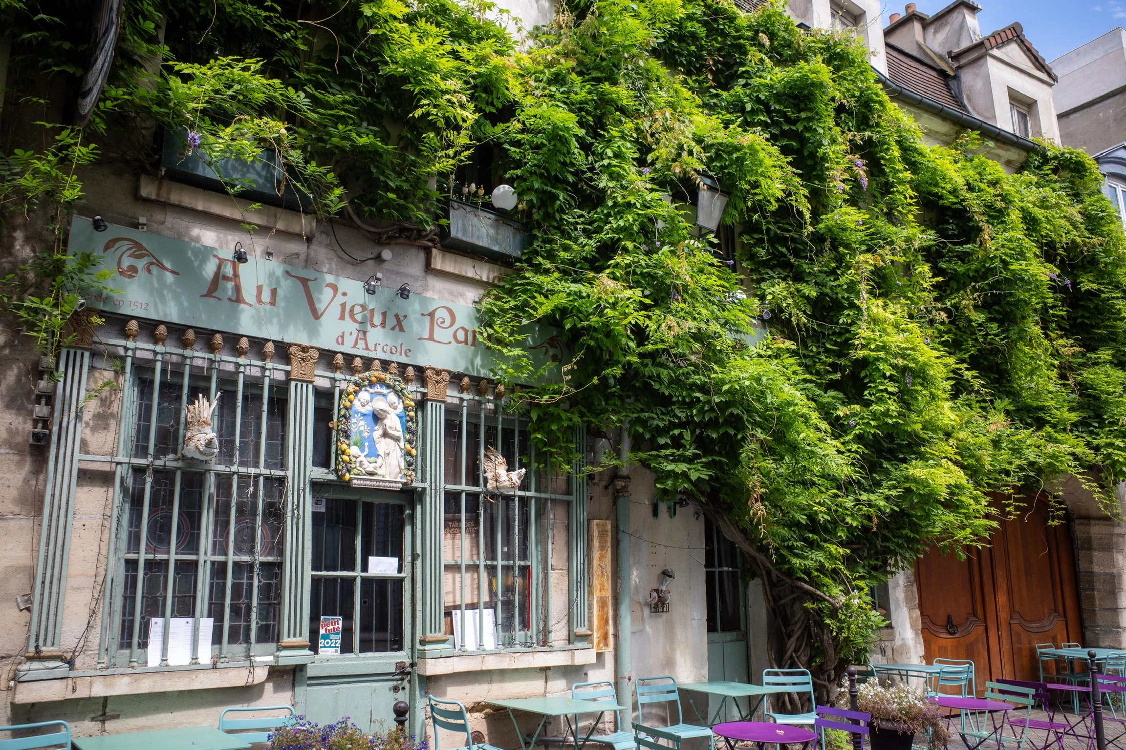 Außenansicht eines kleinen Cafés oder Restaurants, bewachsen mit dichtem grünen Efeu. Vor dem Gebäude stehen bunte Tische und Stühle.