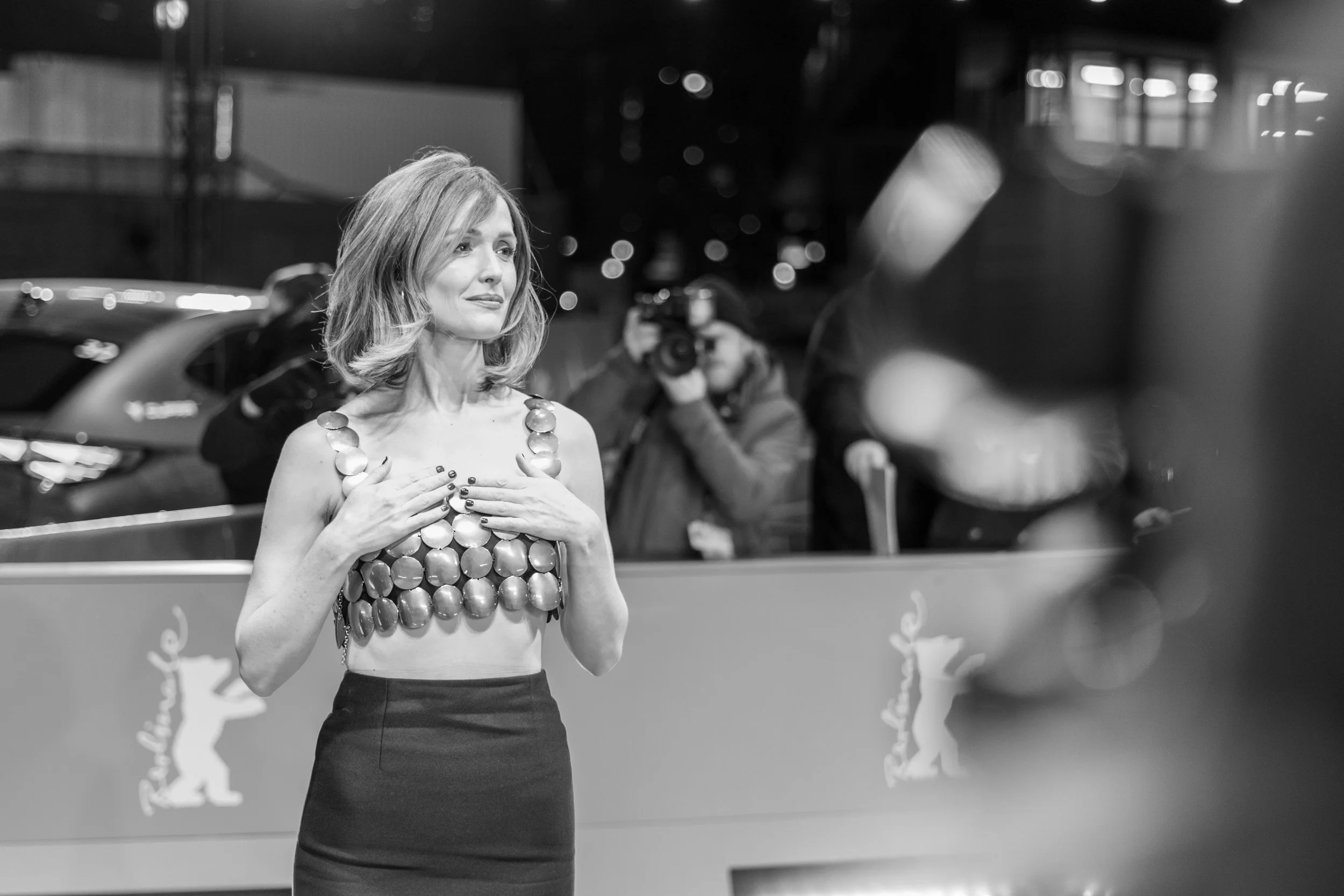 Schwarz-weiß-Foto einer Frau in einem speziellen Kleid aus metallischen Scheiben, umgeben von Fotografen bei einem Event.