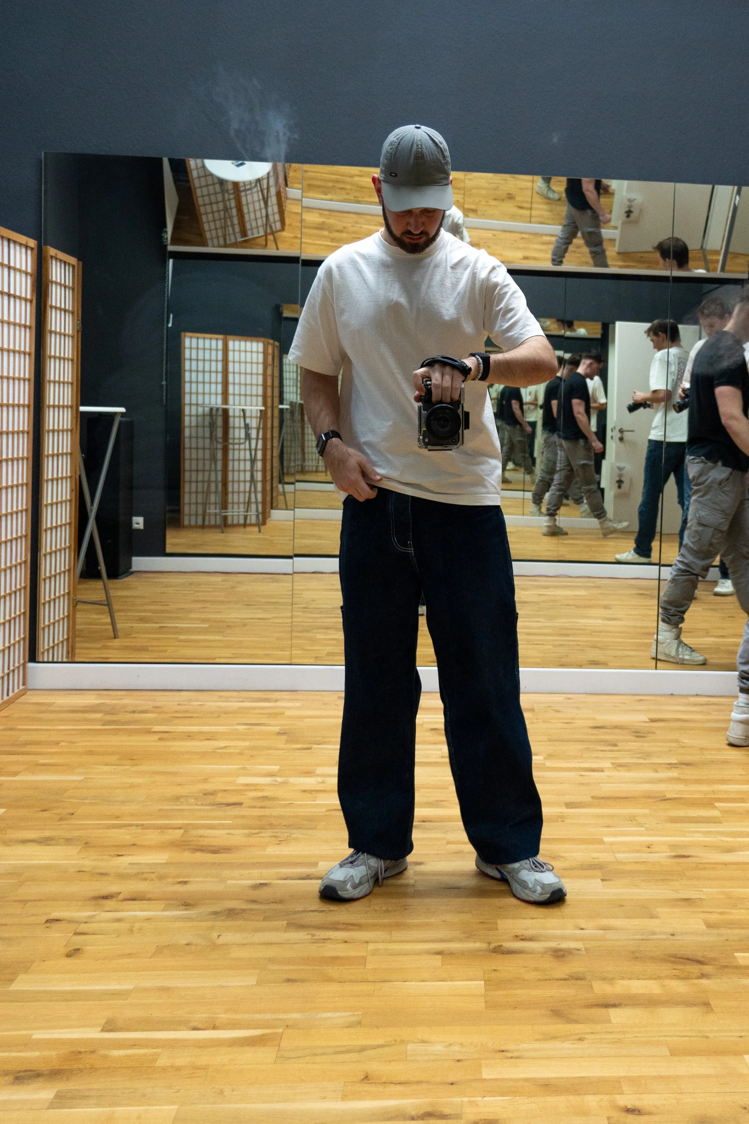 Person macht Spiegel-Selfie in einem Raum mit Holzboden, trägt weiße Mütze, T-Shirt, Jeans und Sneaker, mehrere Spiegel an der Wand reflektieren das Bild.