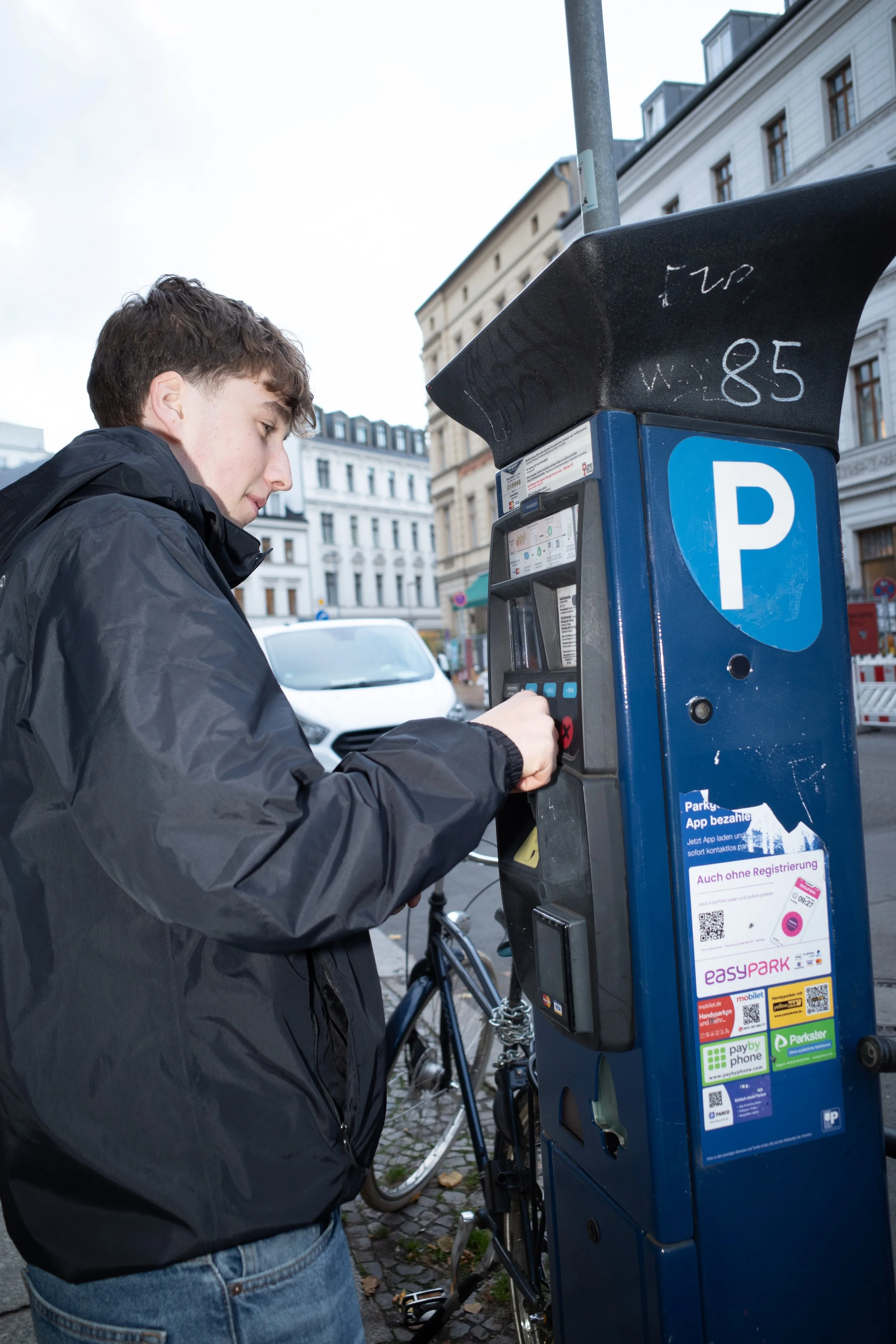 Eine Person, die an einem Parkscheinautomaten steht und eine Münze einwirft, um ein Ticket zu bezahlen. Im Hintergrund sind Gebäude und ein geparktes Auto zu sehen.
