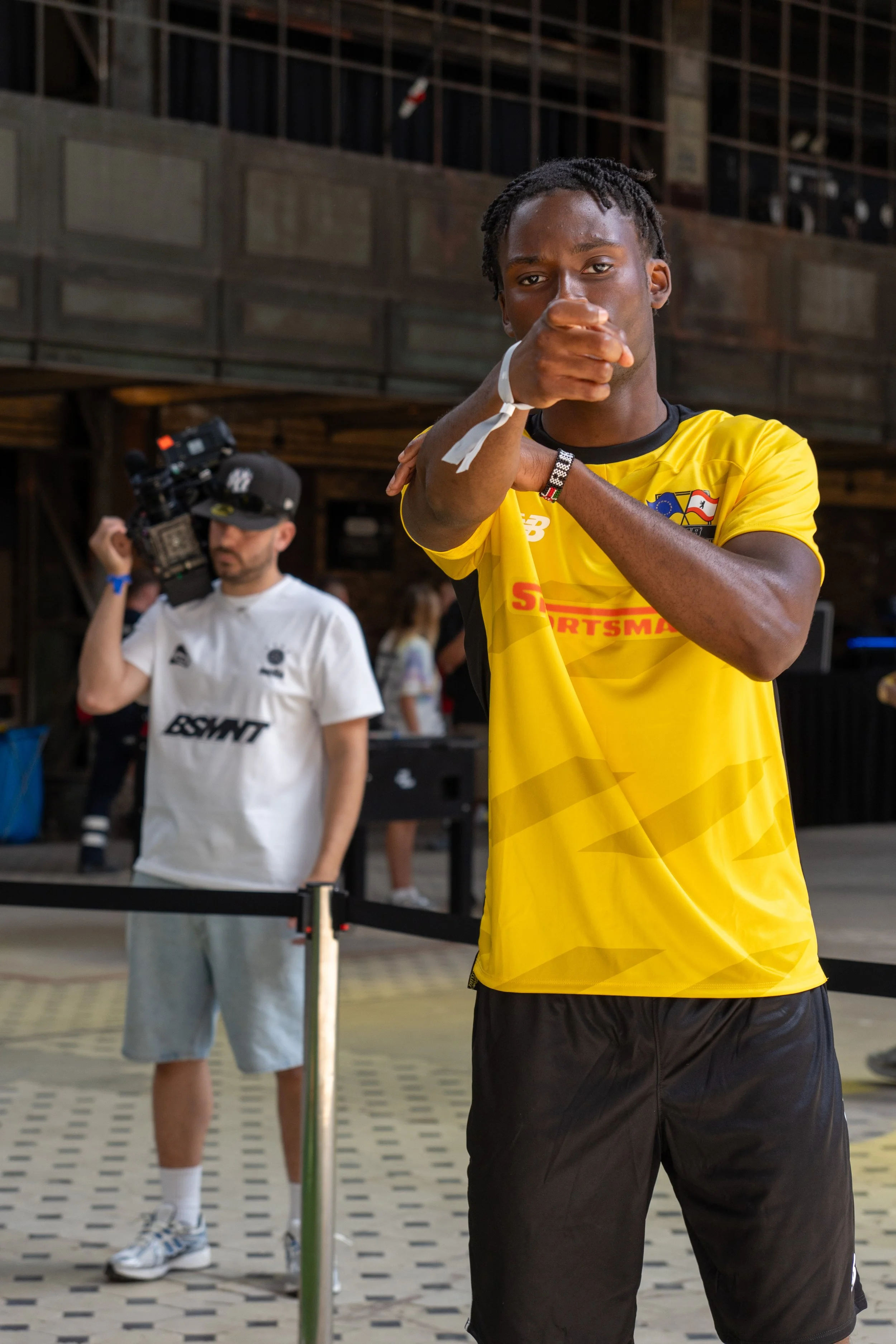 Ein junger Mann in einem gelben Trikot steht vor der Kamera und macht eine Geste mit seinen Armen. Im Hintergrund filmt eine andere Person mit einer Kamera. Die Umgebung scheint eine Halle oder große Innenfläche zu sein.