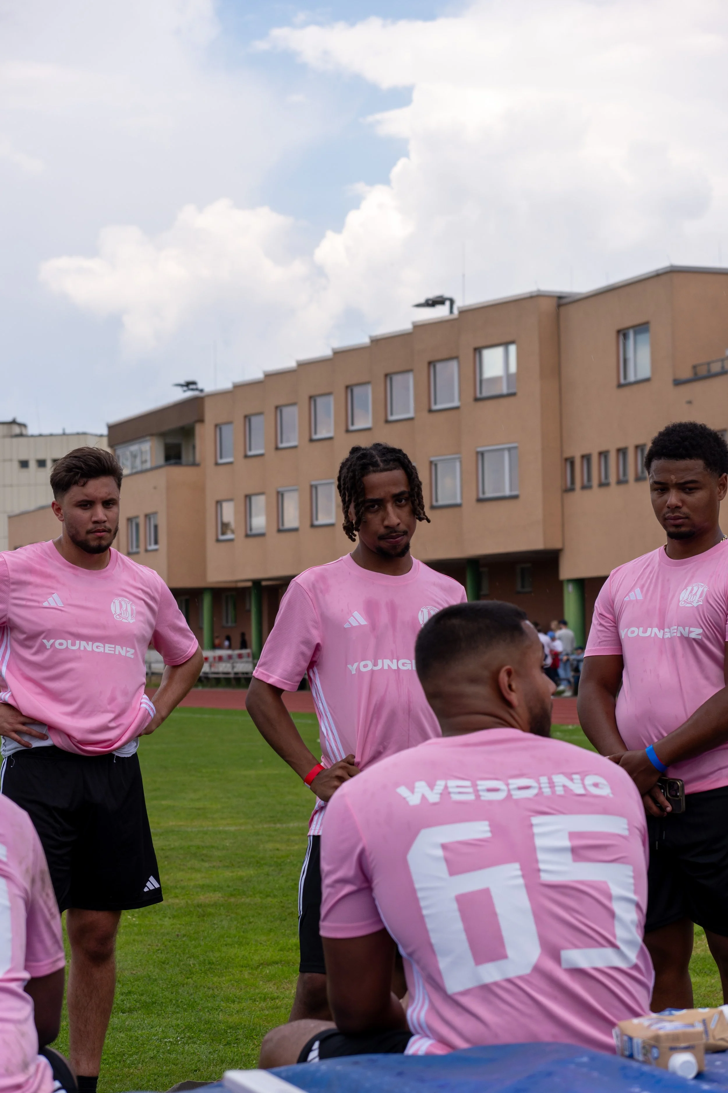 Eine Gruppe von Männern in rosa Fußballtrikots steht auf einem Sportplatz. Einige von ihnen haben ihre Hände in die Hüften gestützt. Im Hintergrund sieht man ein Gebäude mit mehreren Fenstern und Wolken am Himmel.