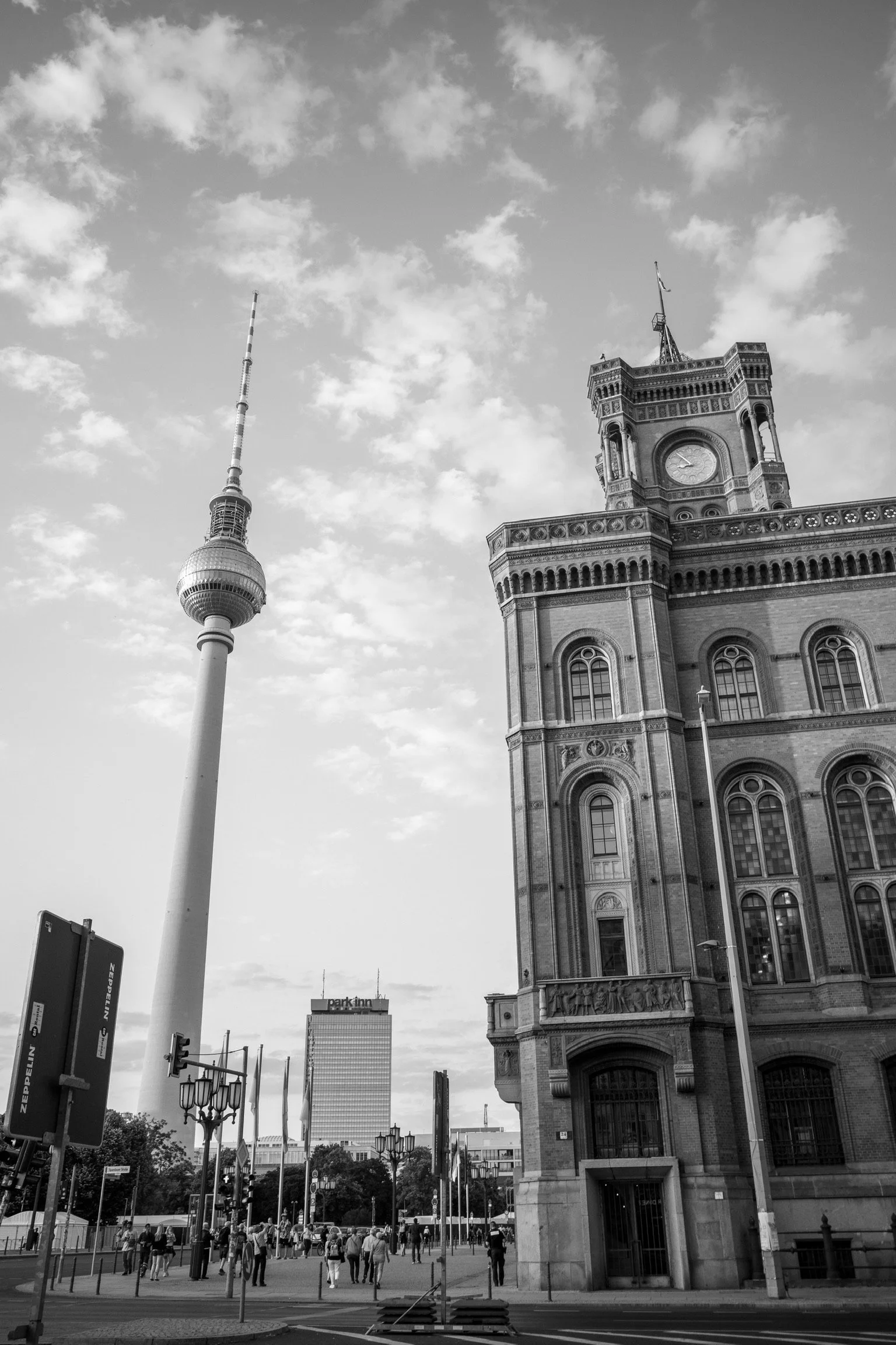 Schwarzweiß-Foto vom Berliner Fernsehturm und einem historischen Gebäude mit Uhrturm, mit belebter Straße und Menschen im Vordergrund.