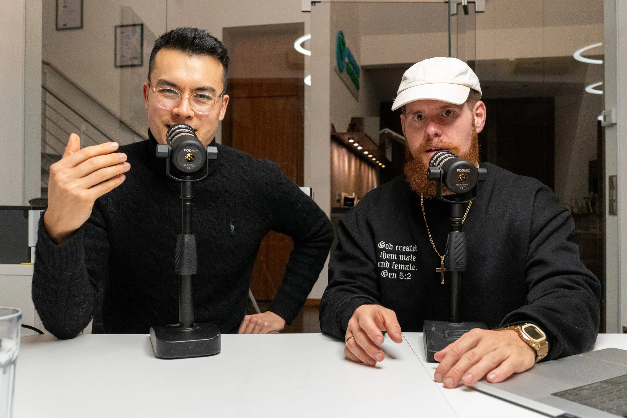 Zwei Männer sitzen an einem Tisch vor Mikrofonen. Einer trägt eine Brille und einen dunklen Pullover, der andere einen hellen Hut und ein schwarzes Sweatshirt. Im Hintergrund befindet sich eine moderne Innenarchitektur.