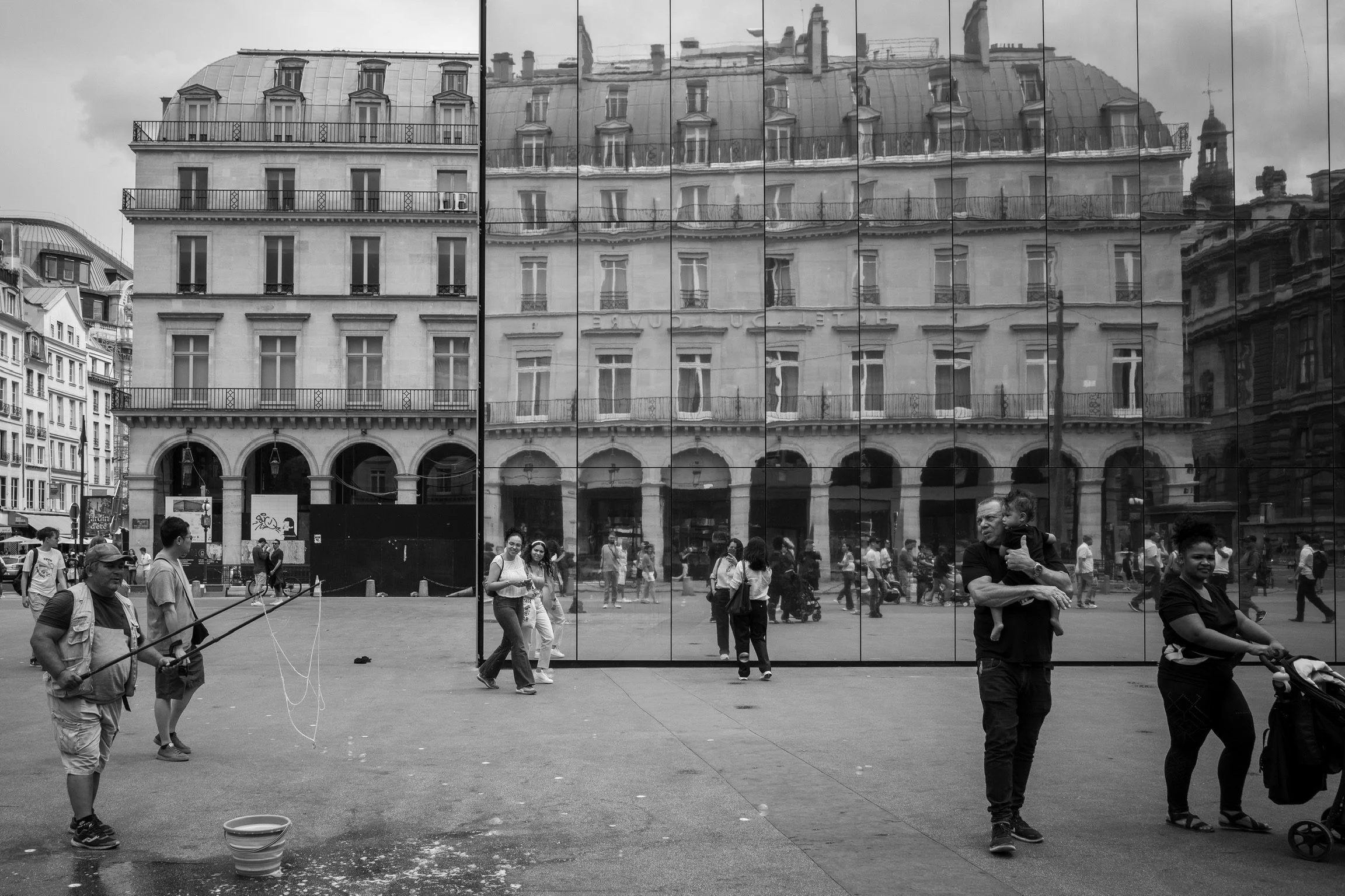 Menschen auf einem öffentlichen Platz mit einem großen, spiegelnden Gebäude im Hintergrund, das die umliegenden Häuser und Passanten reflektiert. Im Vordergrund sind Personen zu sehen, darunter ein Mann, der Seifenblasen macht, und Menschen, die spaz