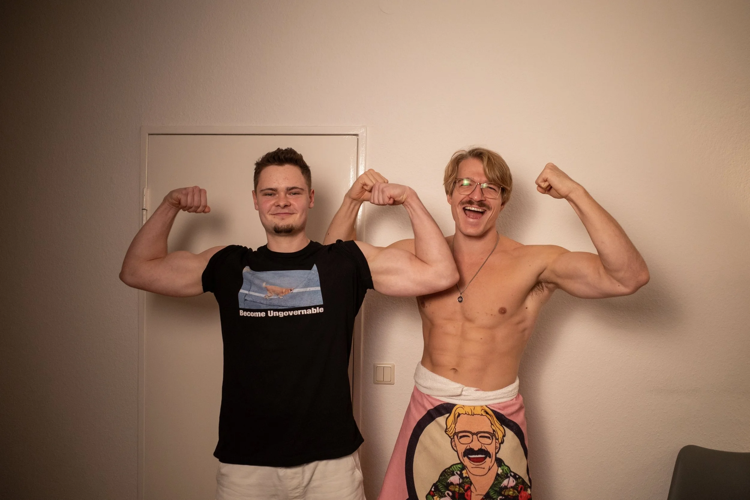 Zwei Männer posieren für ein Foto und zeigen ihre Muskeln. Einer trägt ein schwarzes T-Shirt mit dem Aufdruck "Become Ungovernable" und der andere ist oben ohne, mit einem Handtuch um die Taille. Beide lachen in die Kamera.