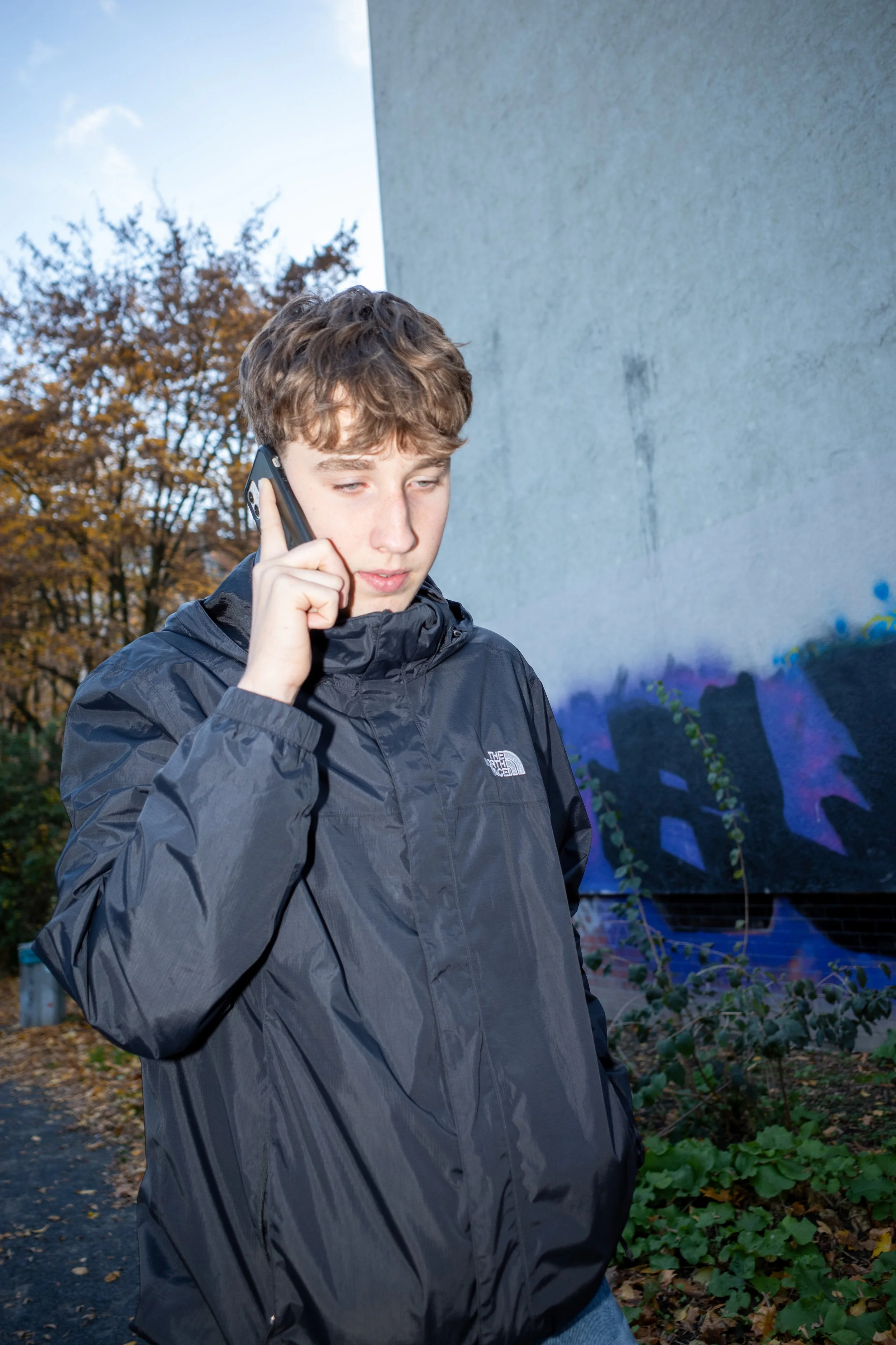 Ein junger Mann in einer dunklen Jacke telefoniert im Freien. Im Hintergrund sind Bäume mit Herbstlaub und eine Wand mit Graffiti zu sehen.