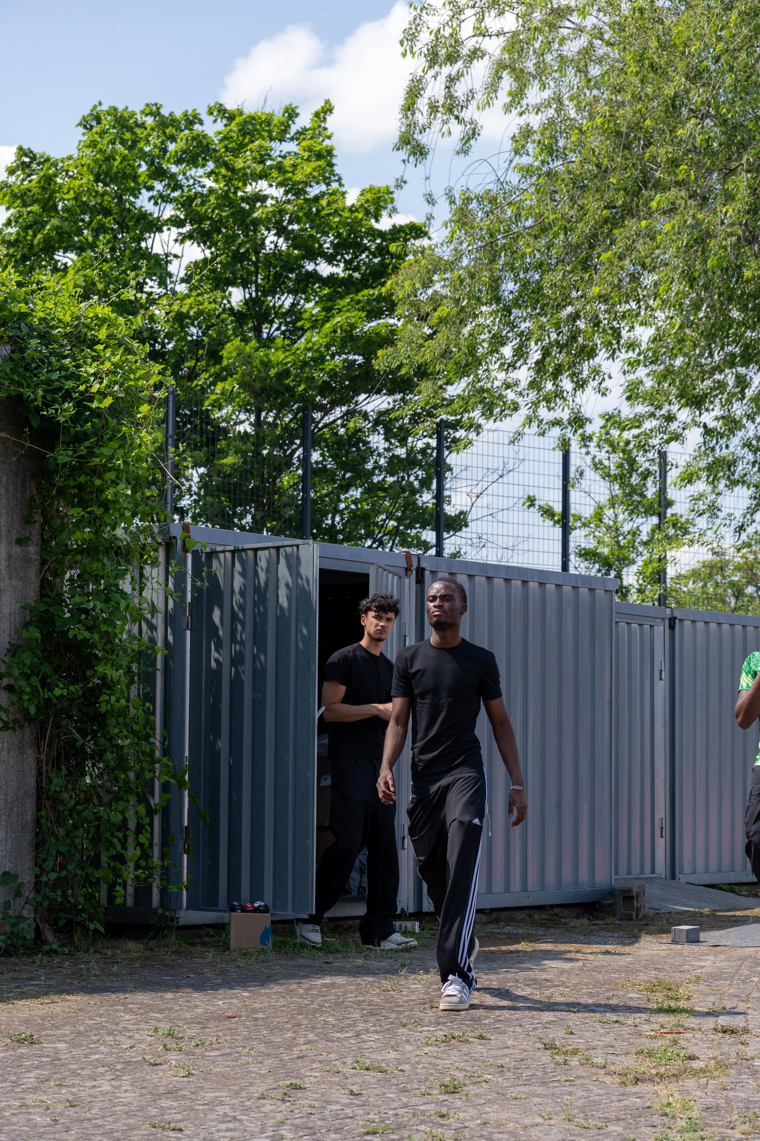 Zwei Männer in schwarzen T-Shirts gehen aus einem grauen Container. Im Hintergrund sind Bäume und ein Zaun zu sehen.
