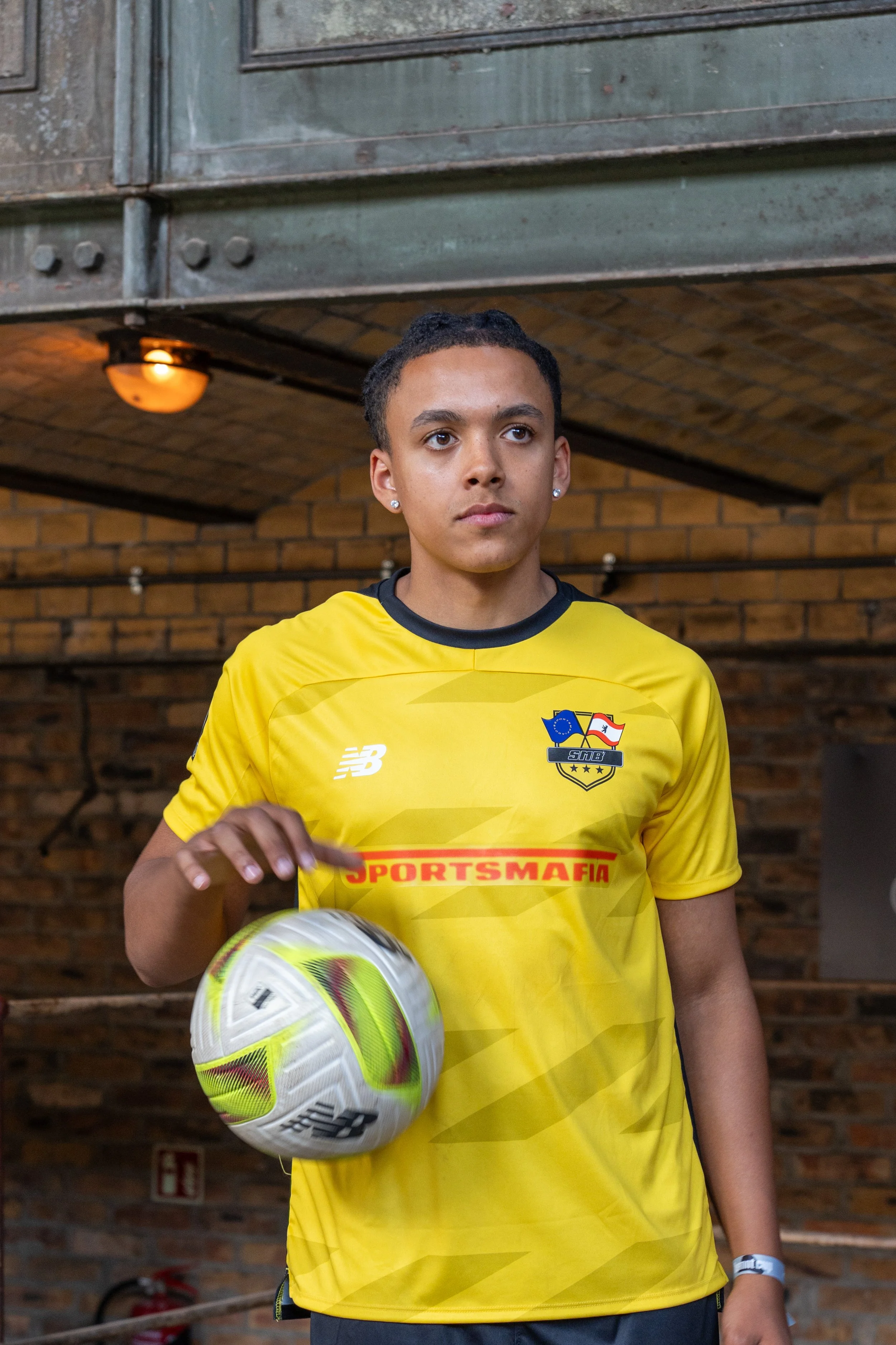 Ein junger Mann in gelbem Fußballtrikot hält einen Fußball in der Hand. Er steht vor einer Backsteinmauer unter einem industriellen Dach.