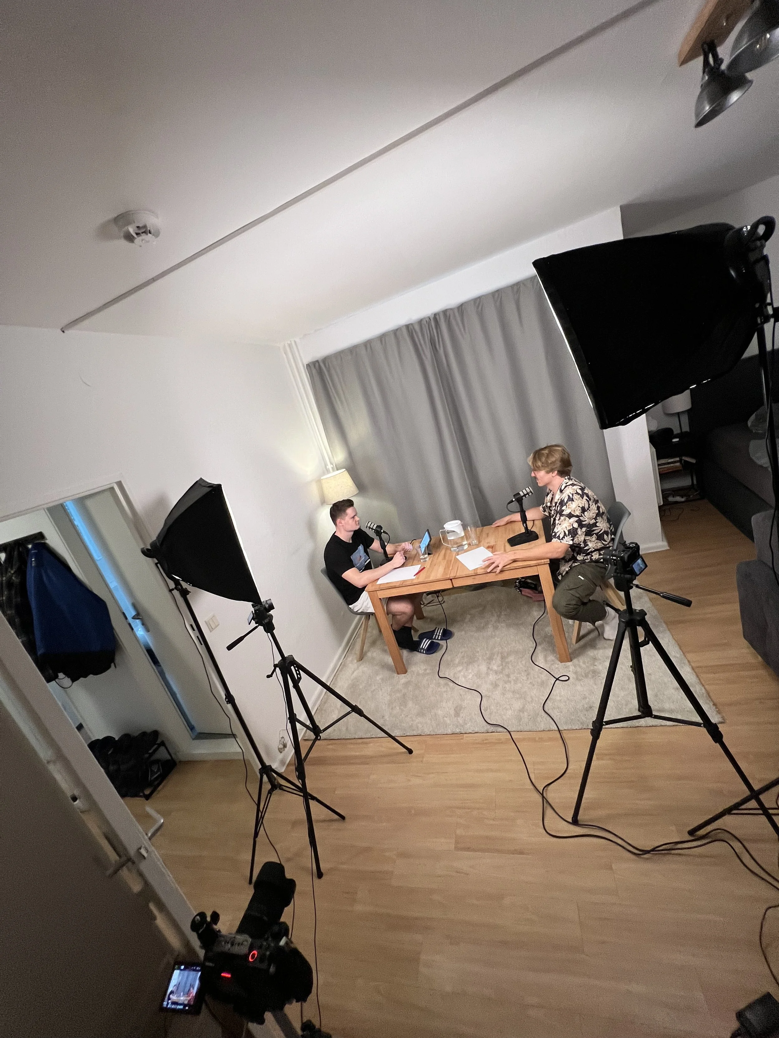 Zwei Personen sitzen an einem Tisch in einem Raum, der für eine Aufnahme vorbereitet ist. Es gibt Mikrofone, Beleuchtungsausrüstung und Kamerastative. Der Raum hat einen Holzboden, eine Lampe und Vorhänge.