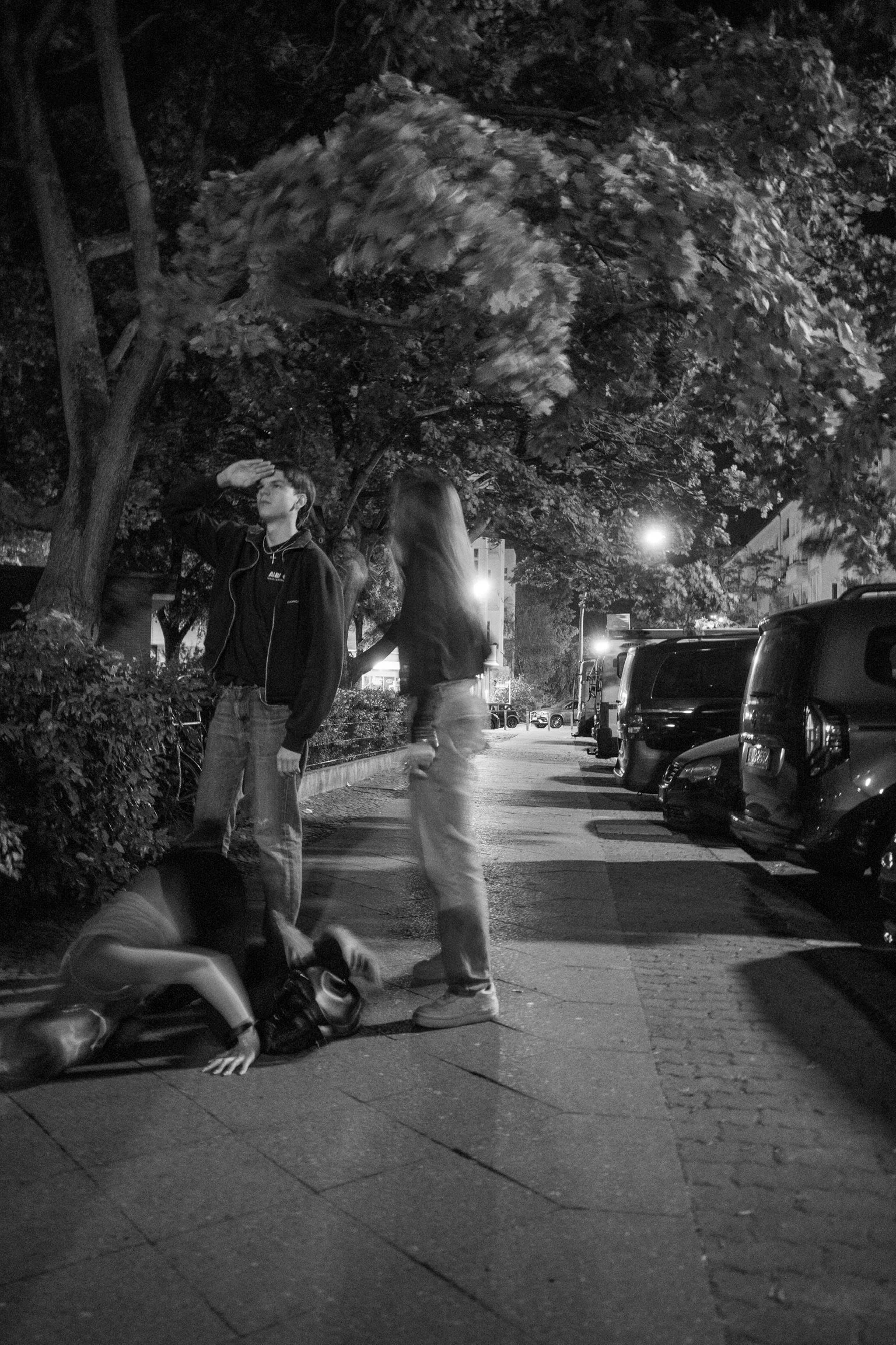 Drei Personen bei Nacht auf einer Straße, eine Person steht, eine steht unscharf, eine kniet am Boden.