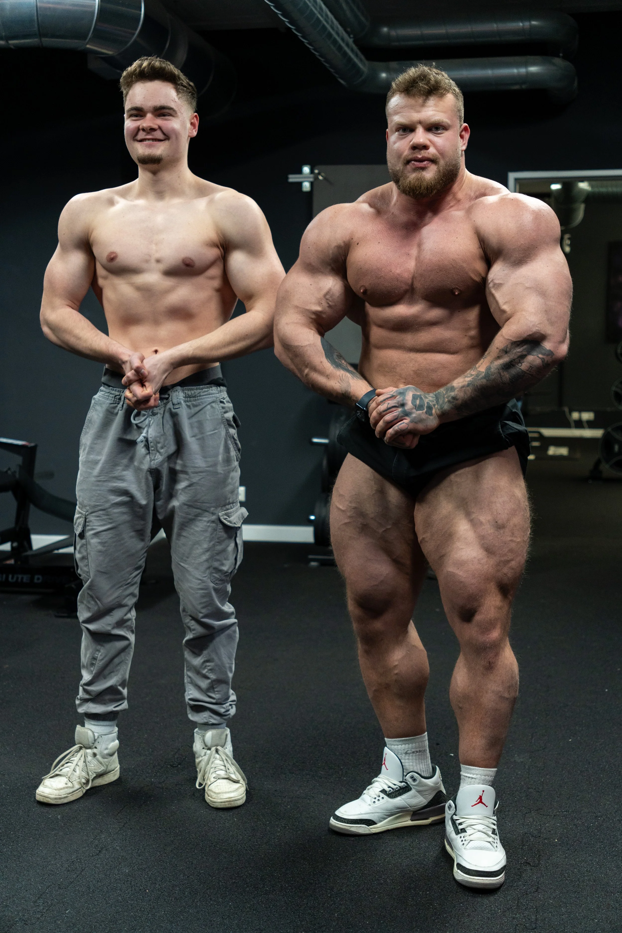 Zwei muskulöse Männer, oben ohne, stehen in einem Fitnessstudio nebeneinander. Der Mann rechts ist beeindruckend muskulös und tätowiert. Beide tragen sportliche Schuhe.