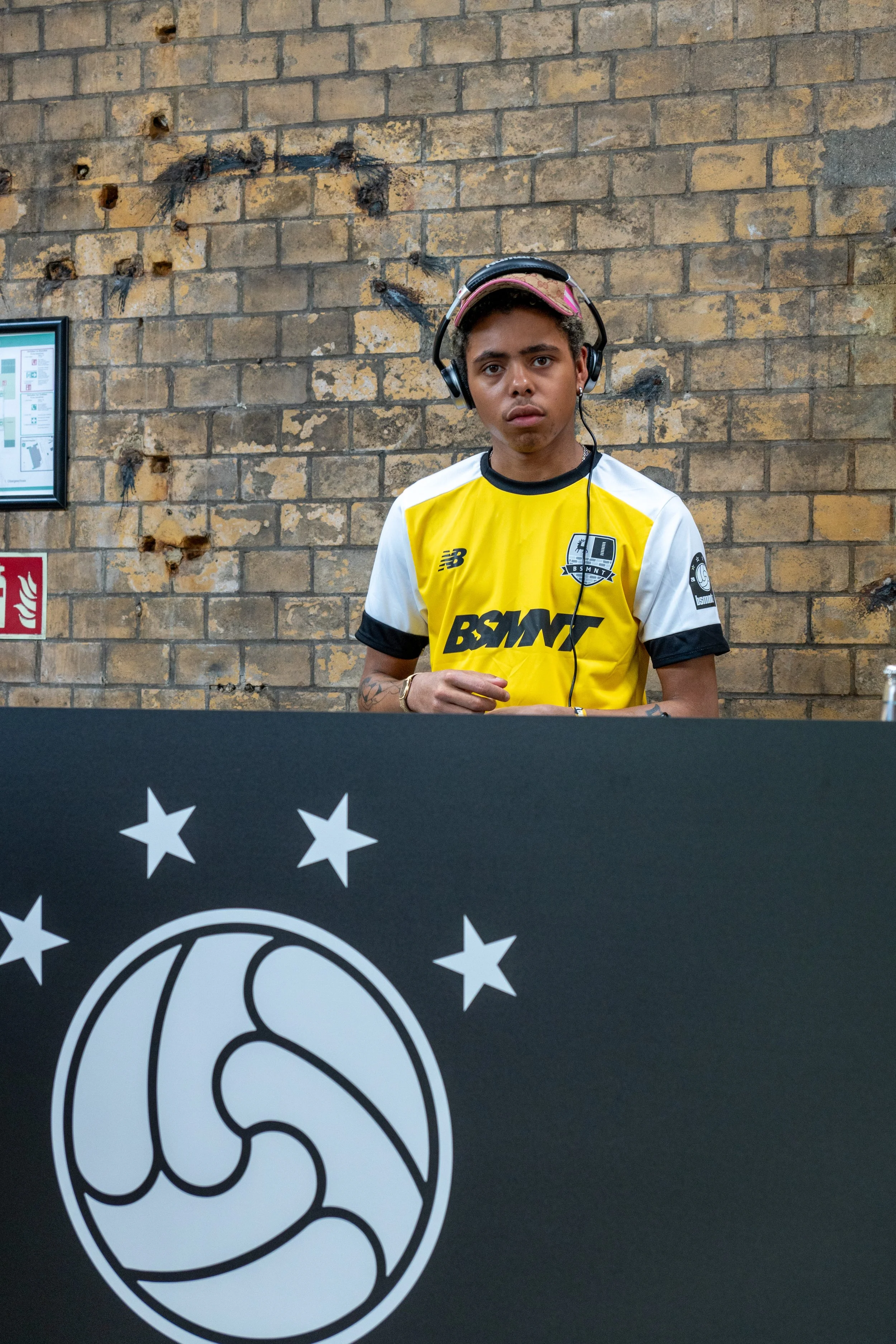 Ein junger Mann mit Kopfhörern steht vor einer Backsteinmauer. Er trägt ein gelb-weißes Trikot mit Aufdrucken unter anderem von New Balance und BSMNT. Vor ihm befindet sich ein schwarzes Pult mit einem emblematischen Kreis und Sternen.
