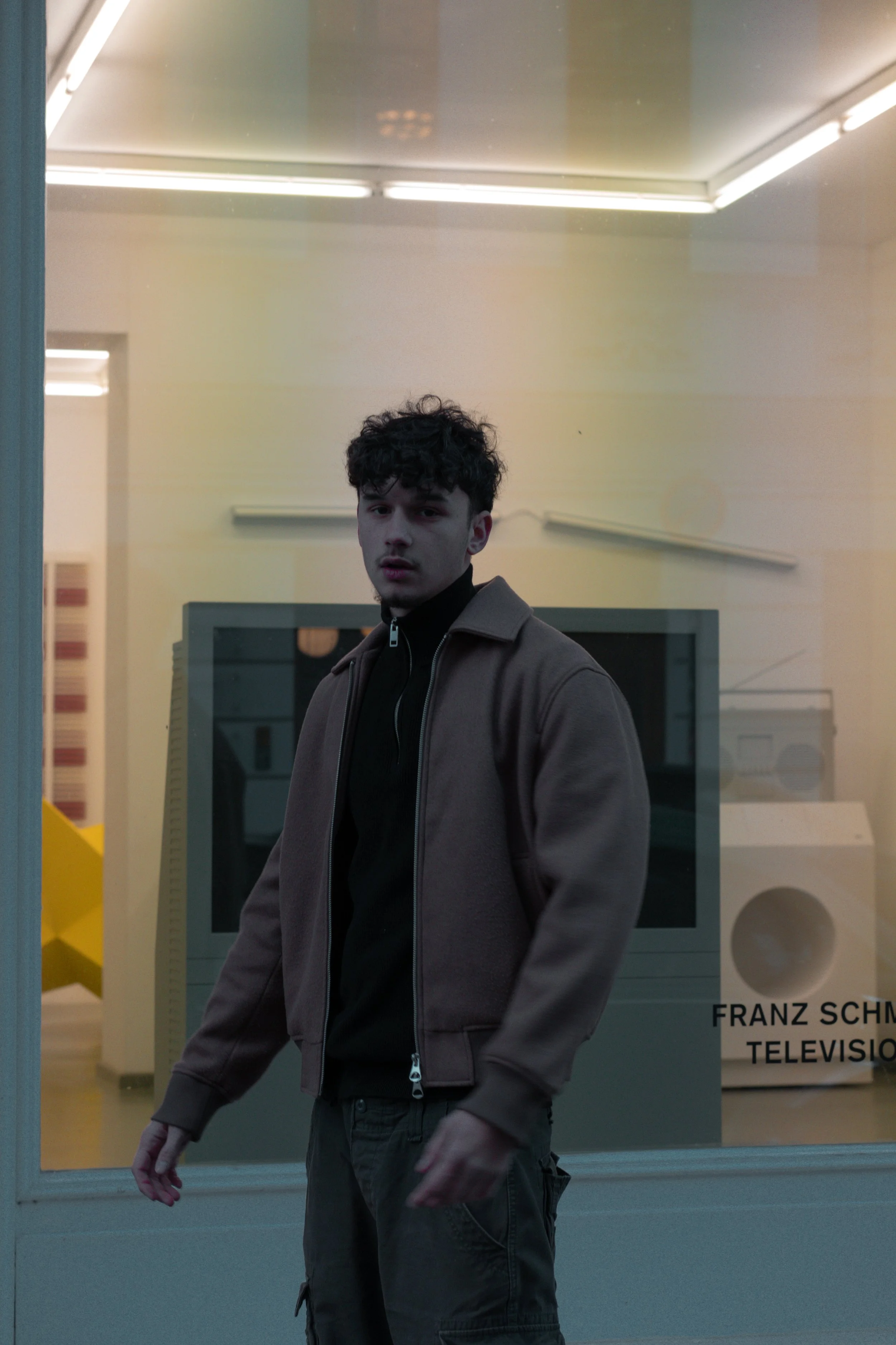 Junger Mann in brauner Jacke steht vor einem Schaufenster mit älterem Fernseher und Lautsprecher.