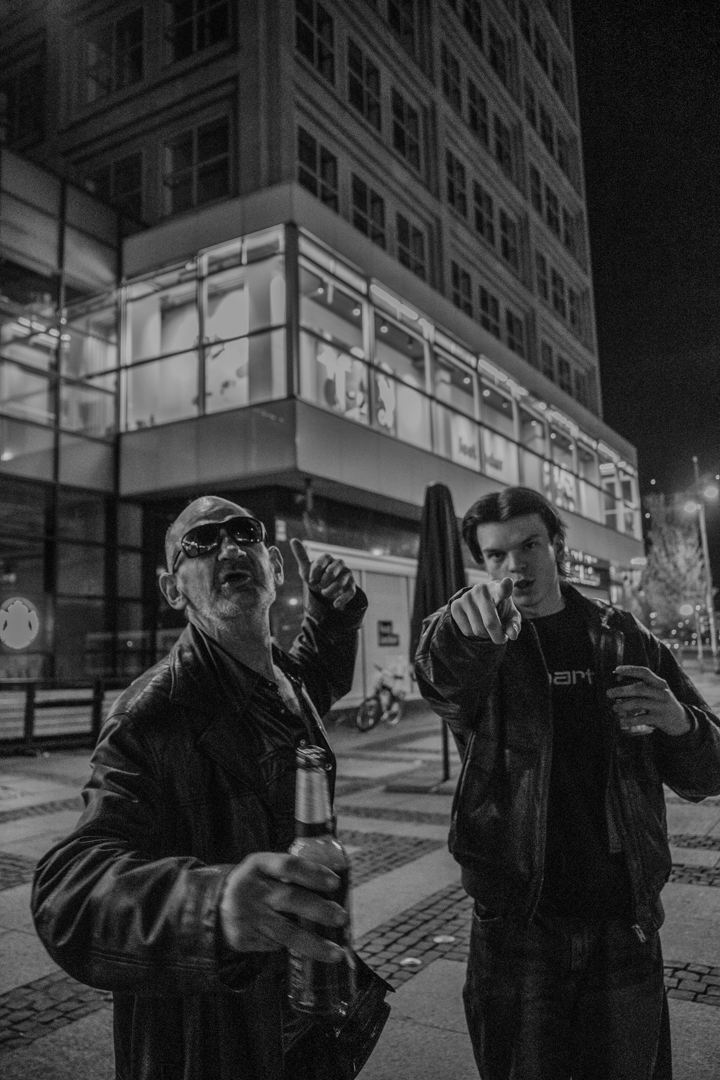 Zwei Männer stehen nachts auf einer Straße vor einem beleuchteten Gebäude. Der ältere Mann trägt Sonnenbrillen und eine Lederjacke, hält eine Flasche und zeigt mit dem Finger. Der jüngere Mann trägt ebenfalls eine Lederjacke, hält ein Getränk in der