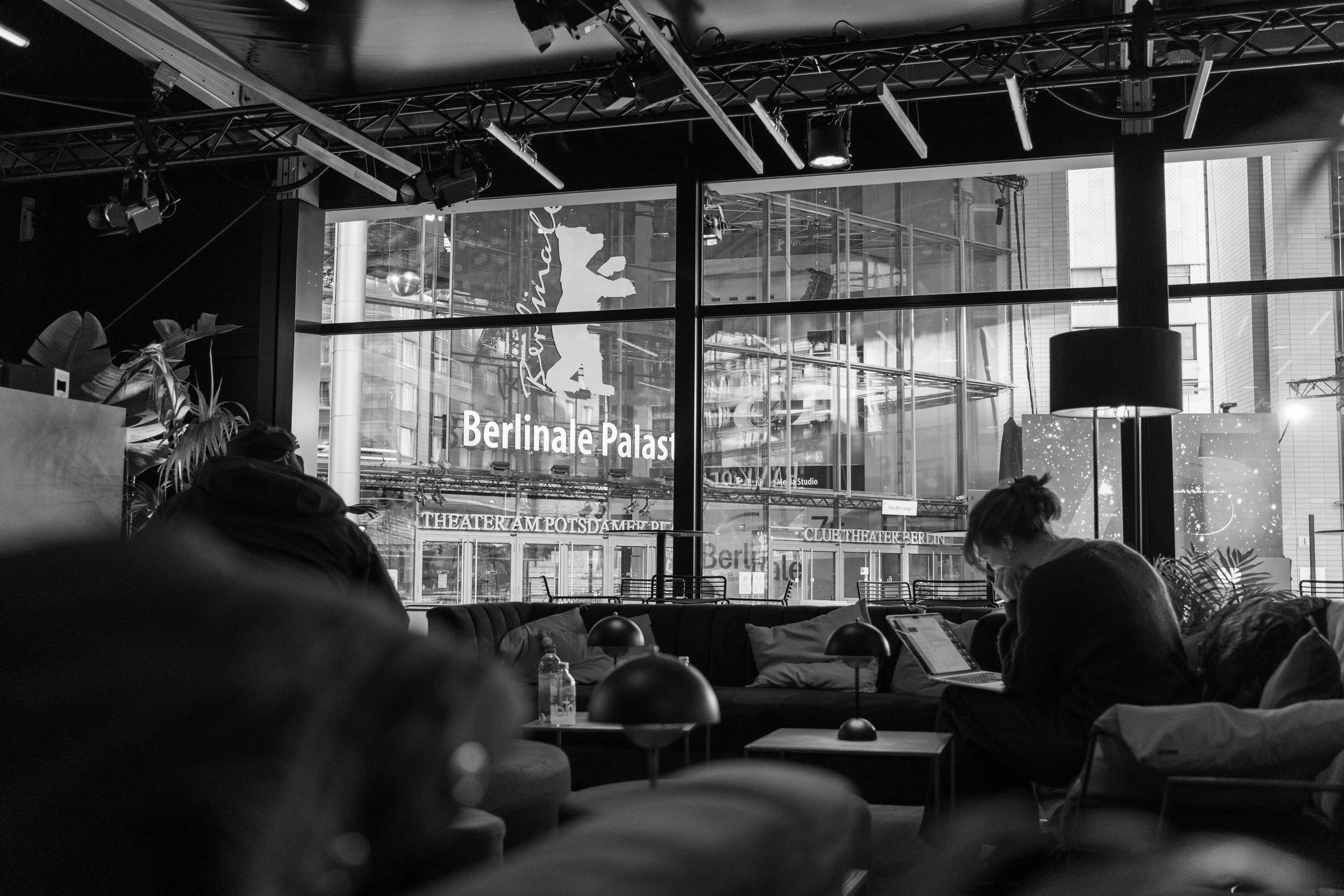 Schwarz-Weiß-Foto von einem modernen Raum mit großen Fenstern, durch die das Logo des Berlinale Palasts zu sehen ist. Im Vordergrund sitzen Menschen an Tischen, möglicherweise in einem Lounge- oder Café-Bereich. Im Hintergrund sind Pflanzen und stilv