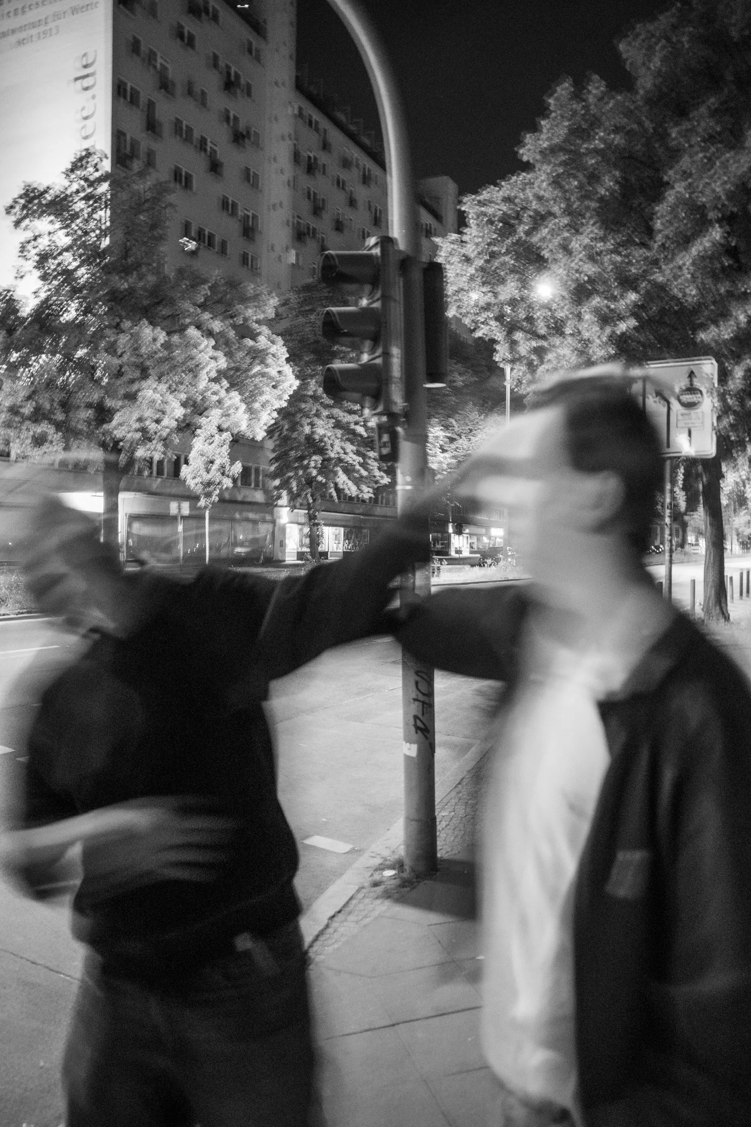 Zwei unscharfe Personen in Bewegung bei Nacht auf einer Straße mit Bäumen, Gebäuden und Ampel im Hintergrund. Schwarz-Weiß-Foto.