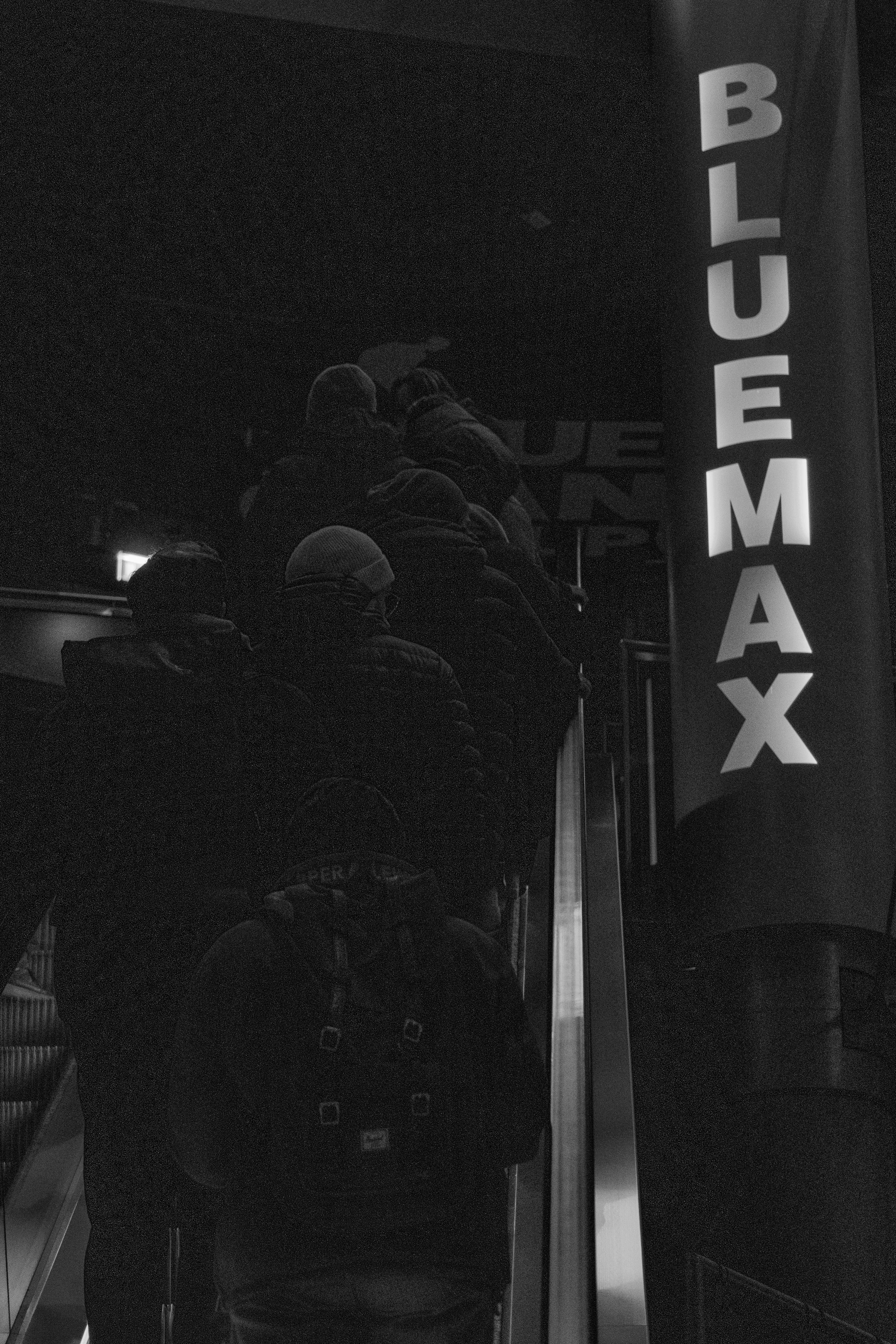 Gruppe von Menschen auf einer Rolltreppe, in einem Gebäude mit "BLUEMAX" Banner.