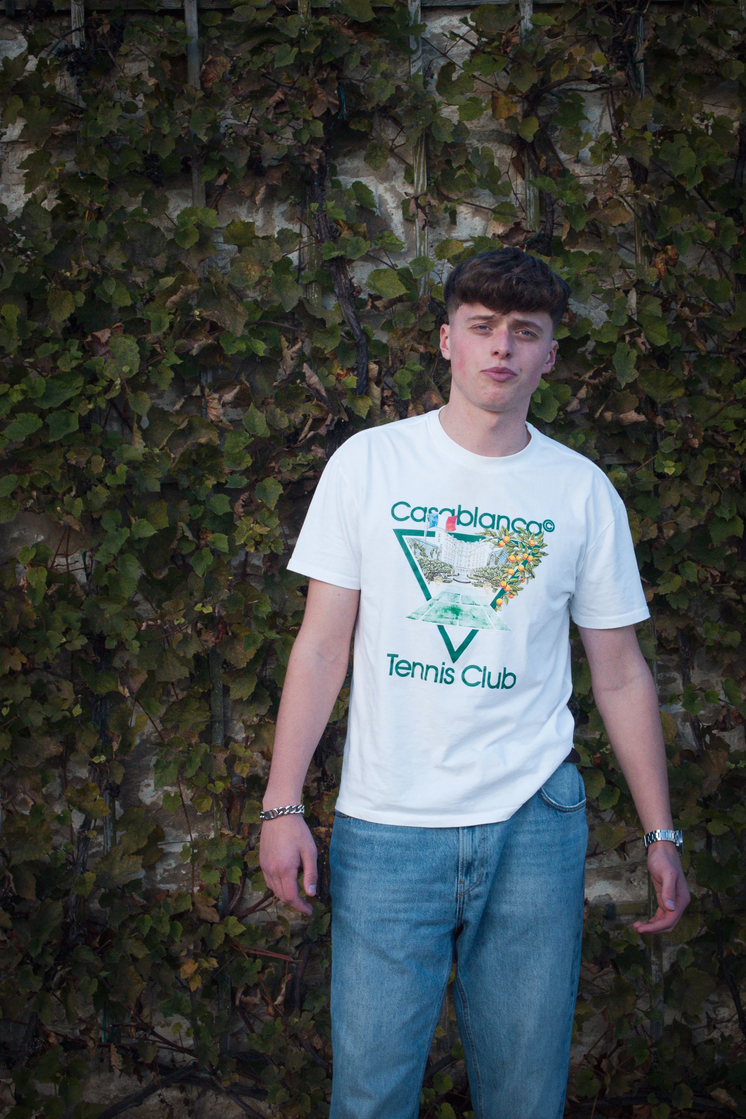 Ein Mann steht vor einer mit Blättern bewachsenen Wand. Er trägt ein weißes T-Shirt mit dem Aufdruck "Casablanca Tennis Club" und eine blaue Jeans.