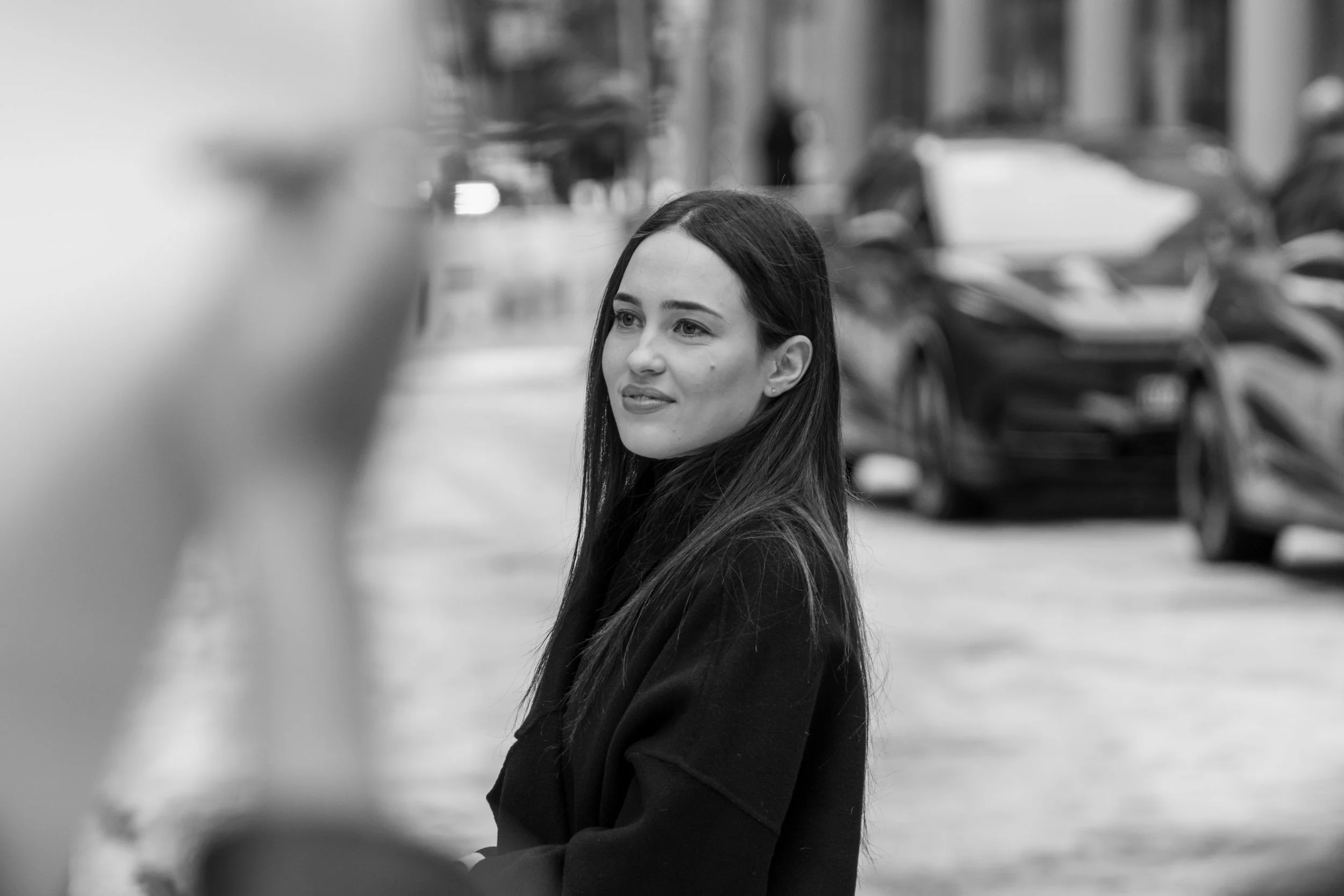 Schwarz-Weiß-Foto einer Frau mit langen Haaren, die in einer städtischen Umgebung lächelt. Im Hintergrund sind unscharfe Autos zu sehen.