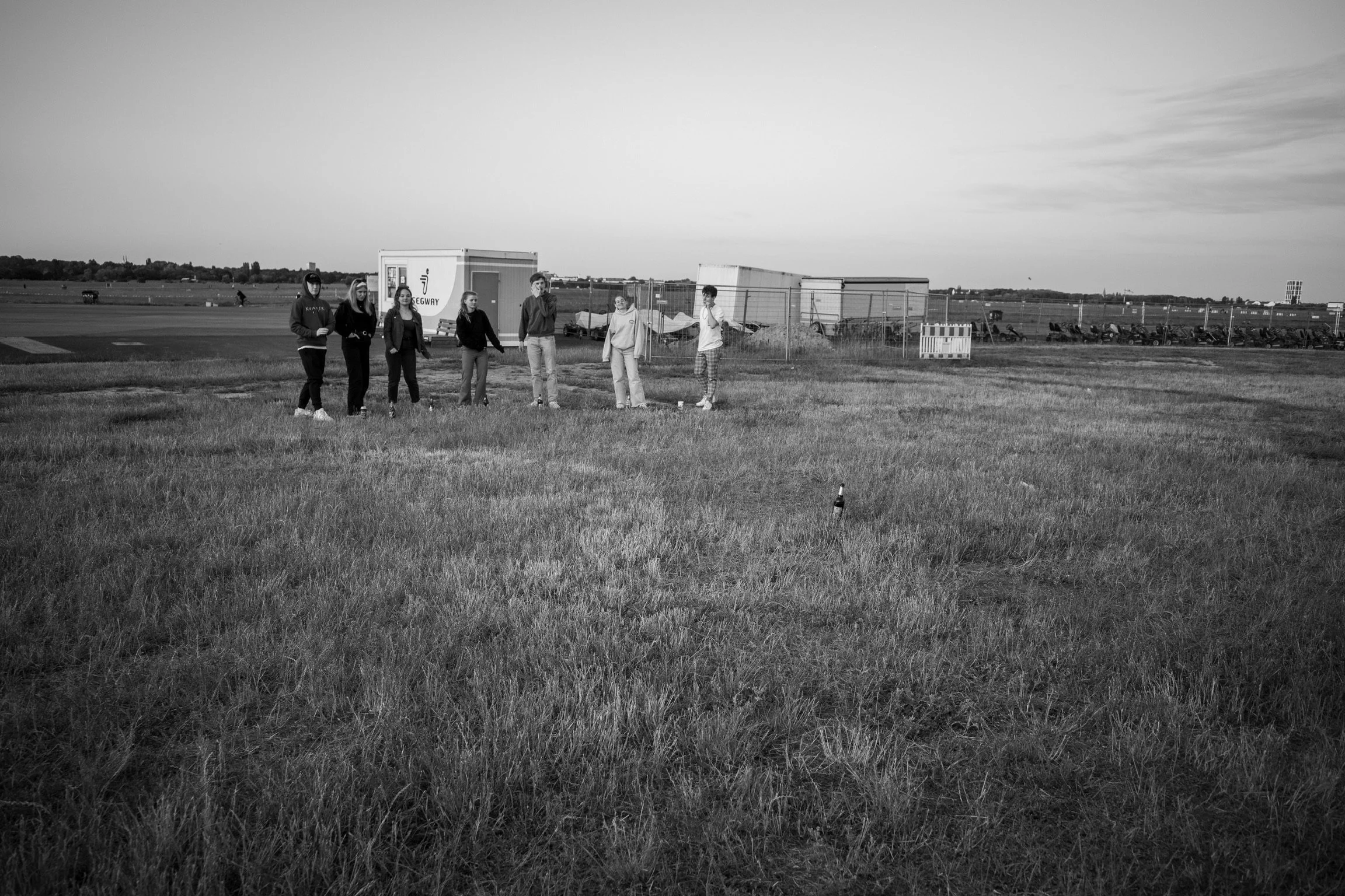 Eine Gruppe von Menschen steht auf einem Feld vor Containern, im Hintergrund sind Bäume und ein Flughafenbereich zu sehen. Eine Bierflasche steht im Gras.