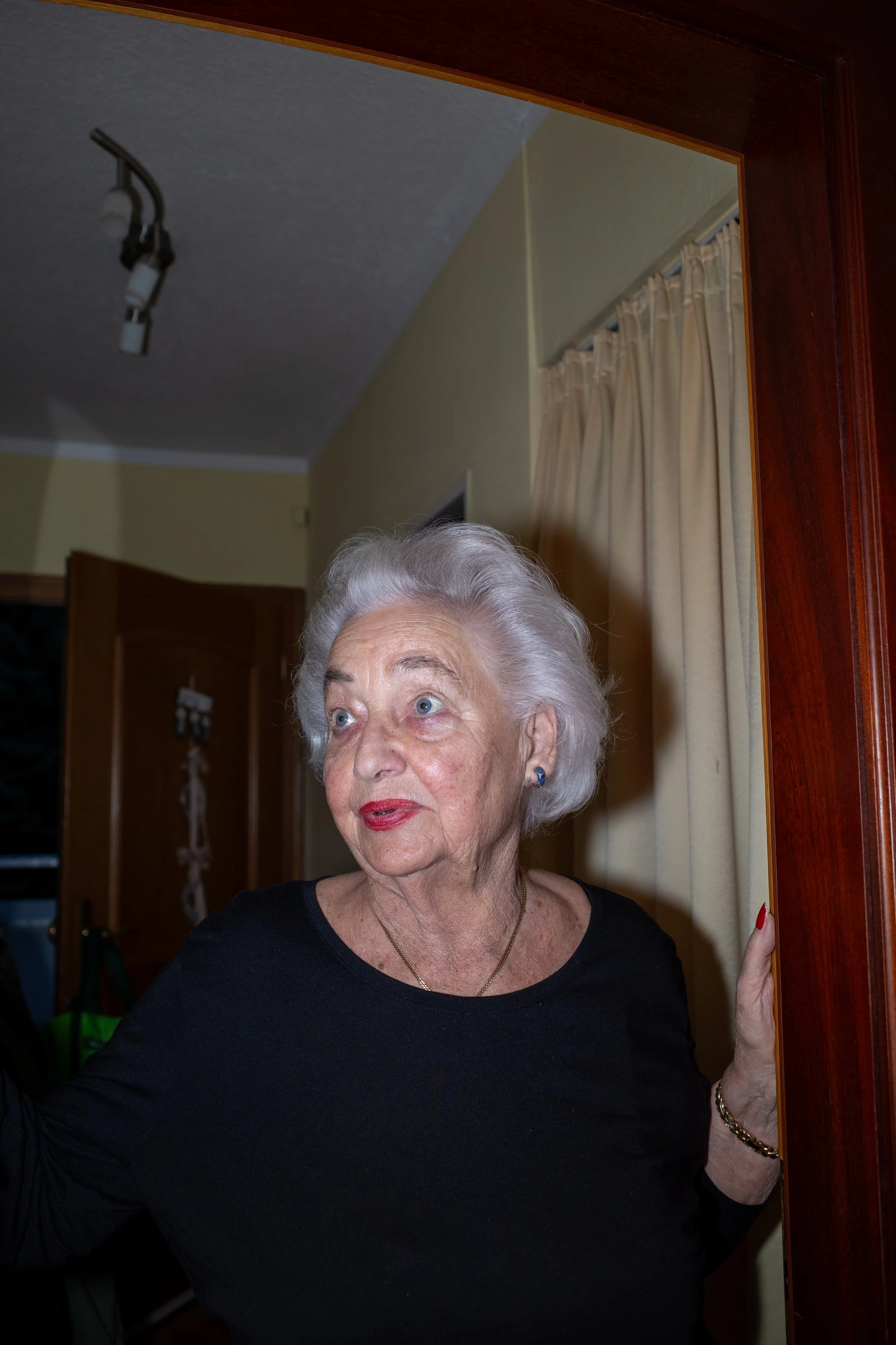Ältere Frau mit weißem Haar steht in einem Türrahmen. Sie trägt einen schwarzen Pullover und ein Armband.