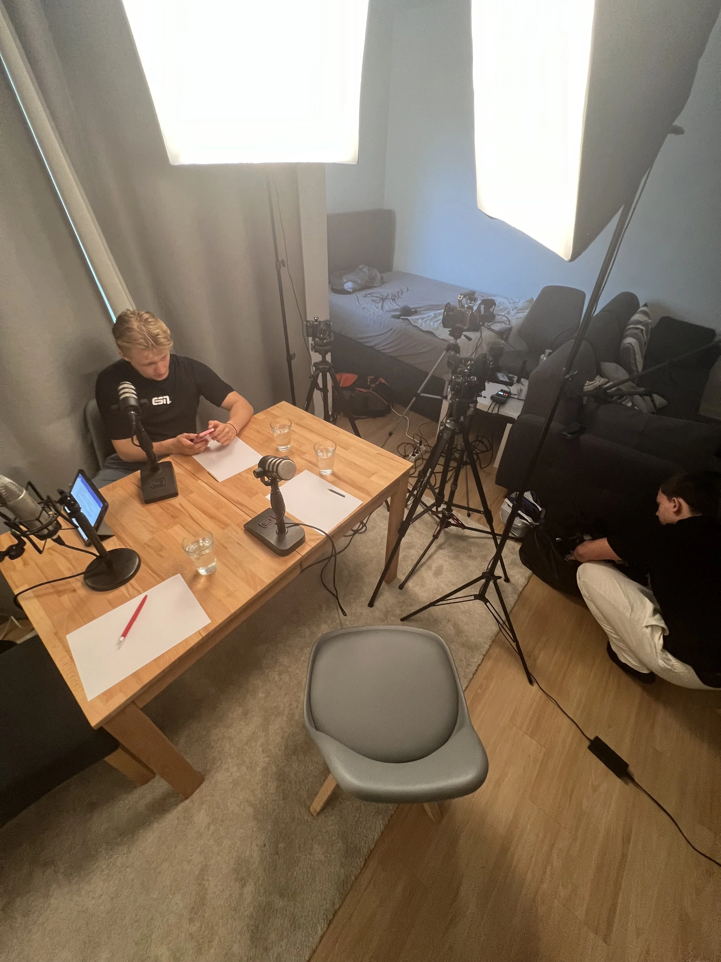 Ein Raum mit einem Tisch, auf dem Mikrofone und Blätter Papier liegen. Eine Person sitzt am Tisch und eine andere Person steht daneben. Der Raum hat professionelle Beleuchtung und Kameras auf Stativen. Im Hintergrund ist ein Bett sichtbar.